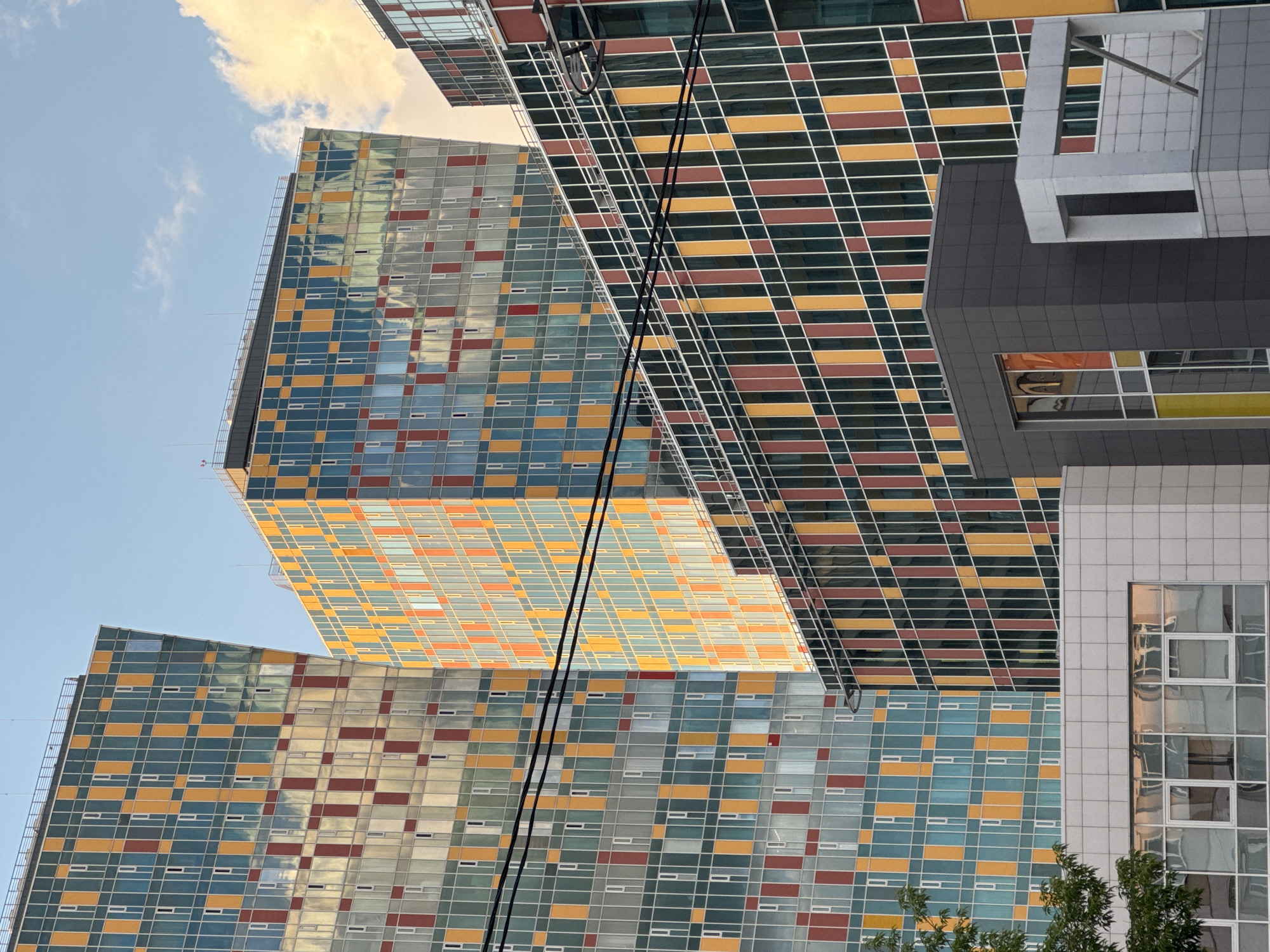 The image presents a striking architectural cityscape in Moscow, Russia, dominated by several modern high-rise buildings under a clear blue sky with scattered clouds. The primary subjects are the tall, glass-clad structures, featuring distinctive, repeating patterns of colorful rectangular panels.
The facades of the main buildings are characterized by a vibrant grid of teal/blue, orange/yellow, and reddish-brown/maroon panels, interspersed with lighter sections. These patterns create a dynamic, mosaic-like appearance across the reflective surfaces. One building on the left exhibits a mix of teal, orange, and white/gray panels, while the large building in the center and to the right displays similar vibrant patterns, with the colors appearing slightly warmer due to direct sunlight.
Power lines, two black wires, cut across the upper central portion of the frame, adding an element of urban infrastructure to the otherwise architectural focus. On the far right, other architectural styles are visible: a building with a dark gray, block-like facade featuring large rectangular windows, and further right, a lighter-colored building with a grid of glass windows. Reflections of the sky and surrounding structures are visible in the glass panels of all buildings.
The scene depicts an urban setting, likely a business or modern residential district, during daytime. The warm light on a prominent cloud and reflections suggest either early morning or late afternoon sun. There are no people visible, and no specific activity is taking place other than the static presence of the buildings. A small amount of green foliage from trees can be seen at the bottom right corner, providing a natural contrast to the artificial structures. No text is visible in the image.