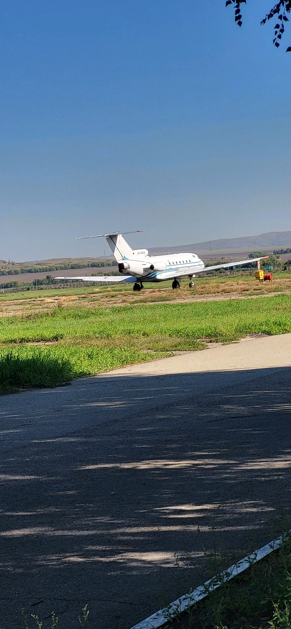 A white jet airplane with blue stripes is parked on a grassy field next to a tarmac. The airplane has two engines under its wings and a single tail fin. The registration number "UR-V405" is visible on the tail. The field is surrounded by a fence and trees in the background, with a clear blue sky above. The tarmac is in the foreground, with shadows cast on it from an unseen object. There are no people visible in the image.