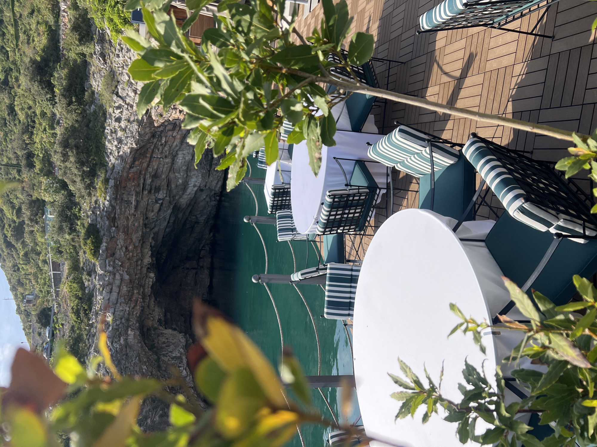 A serene outdoor dining terrace overlooks a picturesque cove in Radovići, Montenegro. In the foreground, several round white tables with white tablecloths are arranged with dark metal chairs. These chairs feature dark green seat cushions and distinctive white and green striped back cushions, neatly placed on a light-colored wooden slatted deck. Lush green foliage, with some branches prominently in the foreground and others softly blurred, frames the scene.
Beyond the dining area, a metal railing system with horizontal ropes separates the terrace from the calm, clear turquoise-green waters of an inlet. The midground and background are dominated by rugged, grey-brown limestone cliffs that plunge into the water, revealing natural caves or overhangs along their base. These cliffs are densely covered with vibrant green Mediterranean vegetation. In the distant background atop the cliffs, a road with parked cars and faint architectural structures can be observed.
The bright sunlight casts distinct shadows from the chairs and railing onto the wooden deck, suggesting a clear day, likely morning or late afternoon. The scene is devoid of people, creating a tranquil and inviting atmosphere ready for dining. No discernible text is visible in the image.