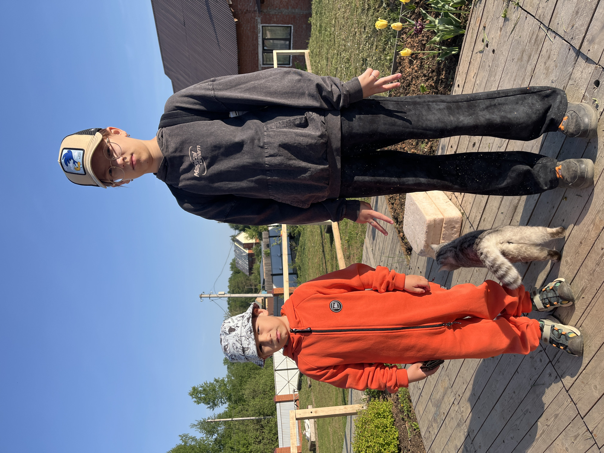 Two children stand on a wooden deck outdoors in Chaikovsky, Russia. The older child wears a baseball cap, glasses, a dark grey hoodie, and dark pants. The younger child wears an orange jumpsuit and a patterned bucket hat. A grey cat rests near the children. The background shows buildings, fences, trees, and a blue sky. Yellow tulips grow in a garden bed.