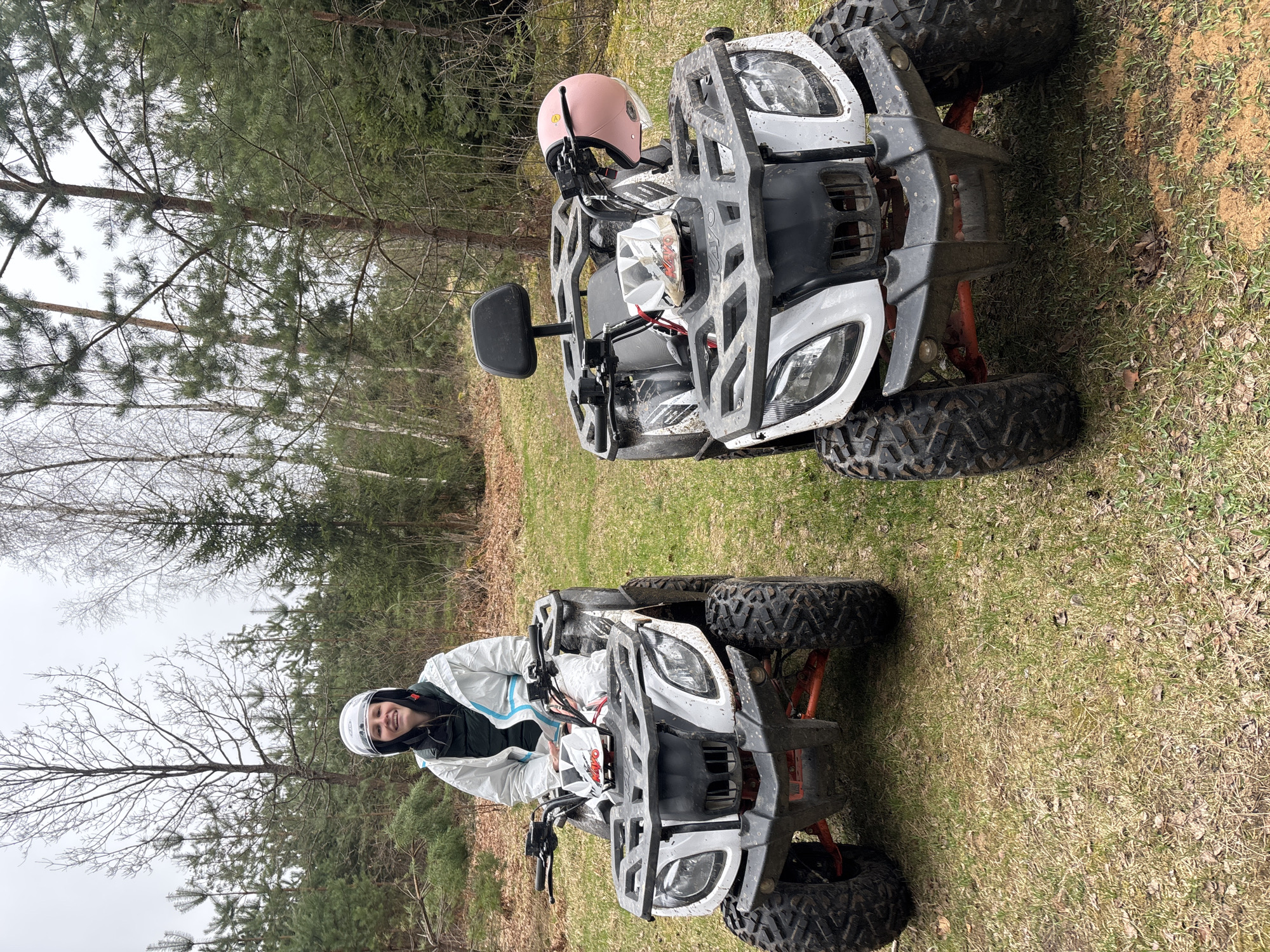 The image depicts two ATVs parked in a grassy area near a forest in Раздолье, Russia. A person wearing a white jacket and helmet is sitting on the ATV in the foreground, smiling at the camera. The second ATV has a pink helmet on its seat. The surrounding environment is characterized by trees and vegetation, suggesting an outdoor setting. The scene suggests a leisure activity.