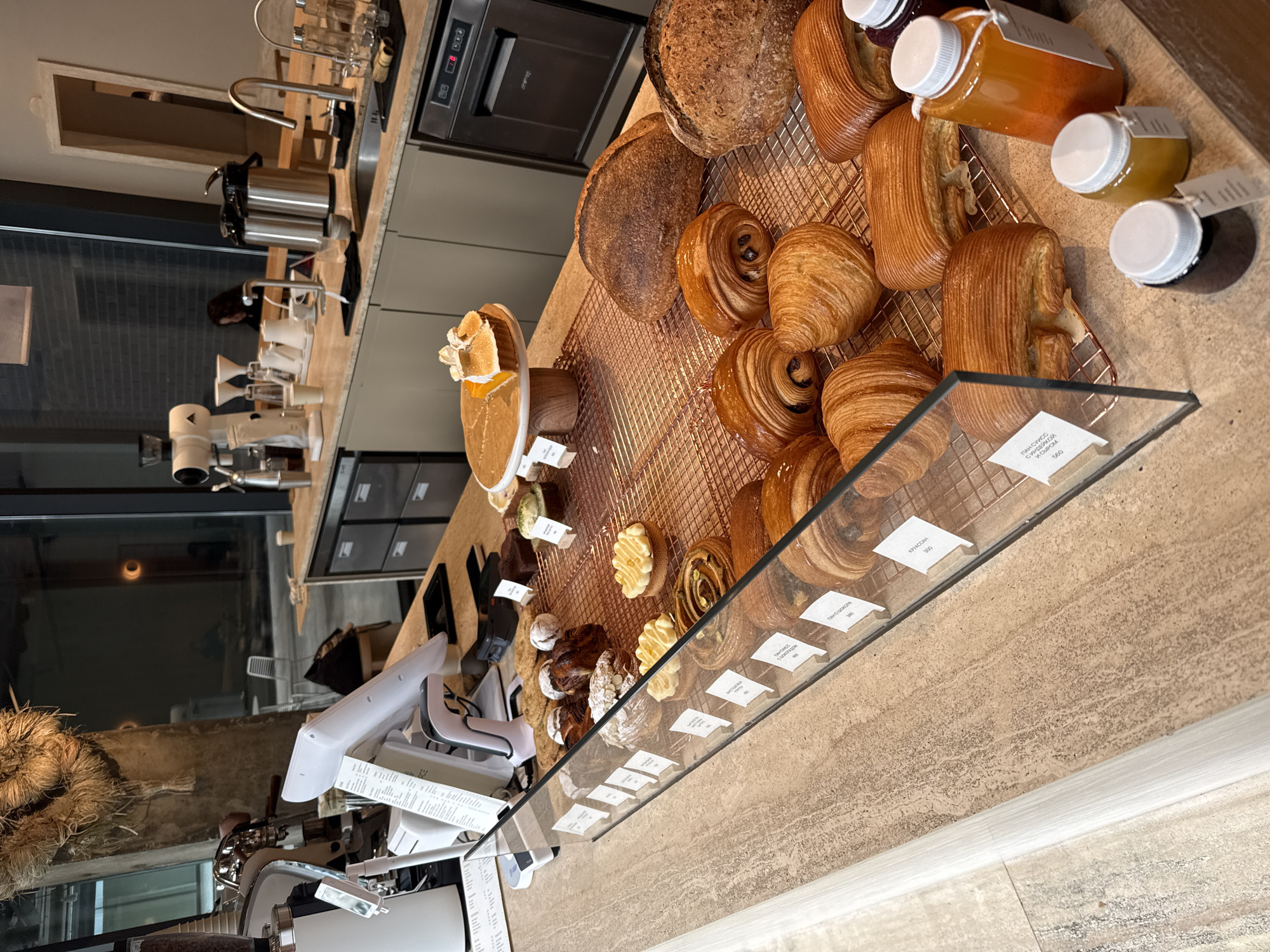 The image depicts a bakery display in Moscow, Russia. Various pastries including croissants, pain au chocolat, and cakes, are arranged on a copper-colored wire rack behind a glass display case. Jars of preserves and loaves of bread are also visible. The display is set on a light brown countertop with a textured surface. A cash register and coffee-making equipment are present on the counter.