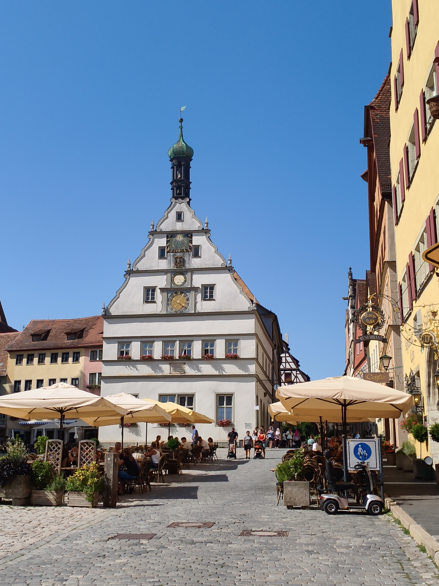 This image depicts a view of the Marktplatz (Market Square) in Rothenburg ob der Tauber, Germany, on a clear, sunny day. In the center of the frame stands the Rathaus (Town Hall), a prominent white building with a distinctive stepped gable. The Rathaus features a green-domed tower topped with a flag and several clocks and astronomical dials on its facade. To the right, a bright yellow building with red shutters lines the square. In the foreground, outdoor seating areas for cafes or restaurants are set up with tables, chairs, and large beige umbrellas. Several patrons are seated at the tables, some appearing to be engaged in conversation. A mobility scooter is parked next to a sign indicating a pedestrian zone. The ground is paved with cobblestones, and potted plants and planters add greenery to the scene. The sky is a vibrant, cloudless blue.