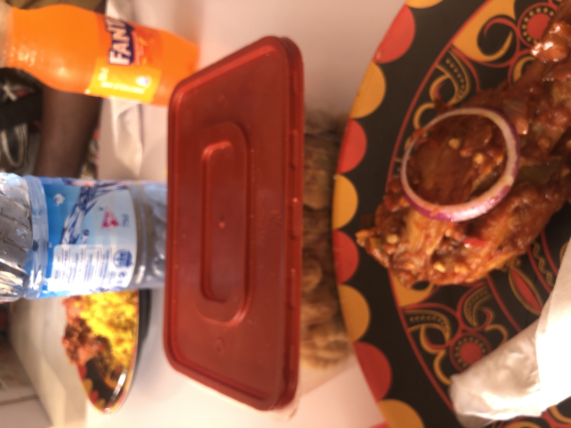 The image shows a table with food and drinks. A bottle of Fanta orange soda and a bottle of water are visible. A red plastic food container is near a plate of food with a red sauce and an onion ring. Another plate with food is partially visible. The location is assumed to be Pindiga, Nigeria, but there is no visual evidence to confirm this. No people are visible. There are no actions or interactions in the image. The environment appears to be indoors.