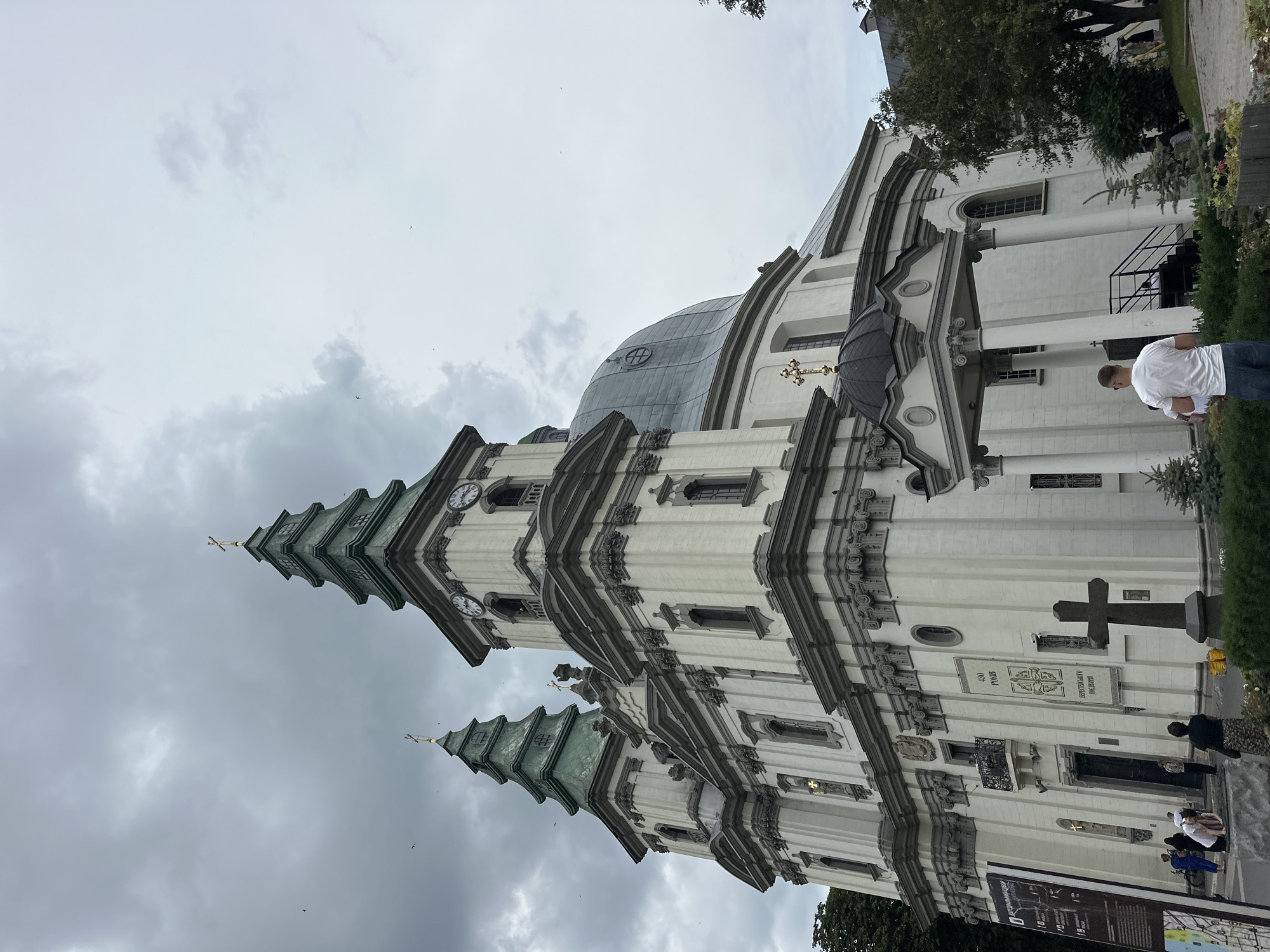 The image shows the Ternopil Cathedral (also known as the Cathedral of the Immaculate Conception of the Blessed Virgin Mary) located in Ternopil, Ukraine. The imposing white stone building features two prominent green-roofed towers, each topped with a golden cross, and a large dome. The façade is adorned with intricate architectural details, including columns, cornices, and decorative moldings. Clocks are visible on one of the towers. Several people are present in the foreground, some interacting with a large stone cross on the left side of the image, while others are walking near the entrance. A man in a white t-shirt and blue shorts is standing with his back to the camera, facing the church. A tall informational board is positioned to the left of the entrance. The sky is overcast with dramatic clouds, and a few birds are flying in the distance. Trees and bushes surround the cathedral.