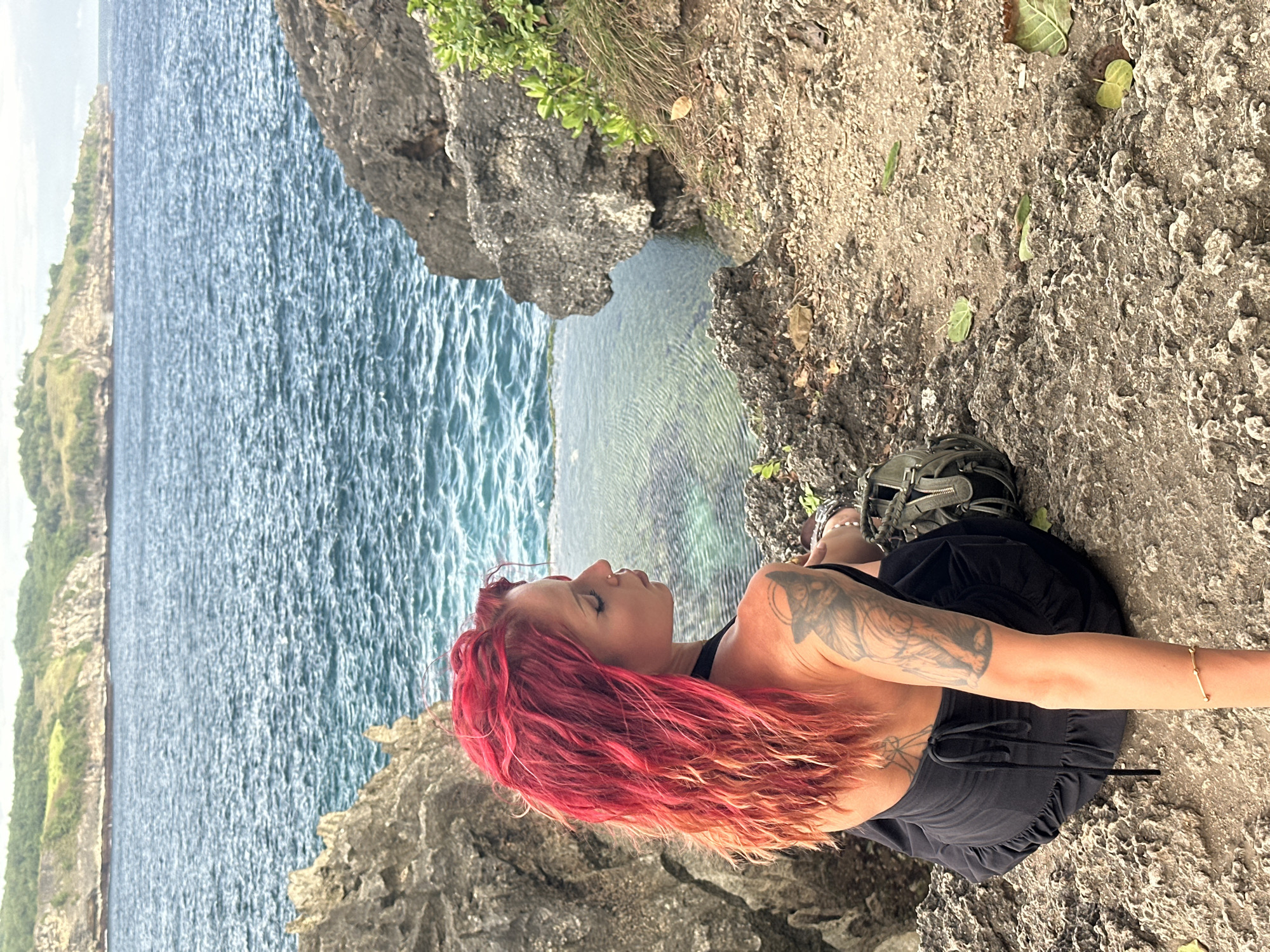 A woman with red and orange hair is sitting on a rocky cliff overlooking the ocean in Nusa Penida, Indonesia. She is wearing a black dress and a gold bracelet. She has tattoos on her back and arm. A pair of green sandals sit near her feet. The water is clear and blue. The rocky cliff is covered with some vegetation.