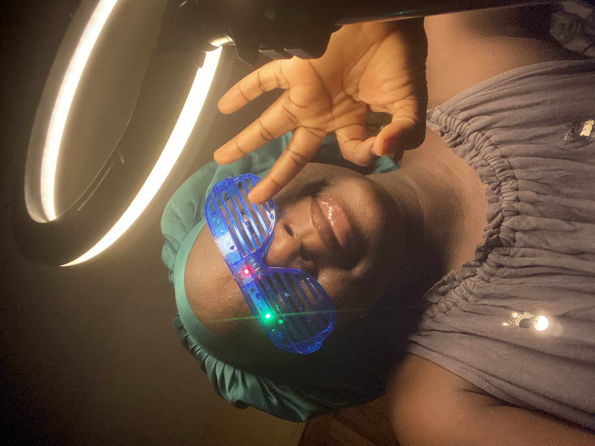 A close-up shot features a person lying down. The individual is wearing blue light-up glasses and a green head covering. A hand is positioned above their face, making a gesture. A ring light is visible in the upper left corner. The person's clothing appears to be a gray, gathered top. No other details suggest a location in Maigizo, Nigeria.