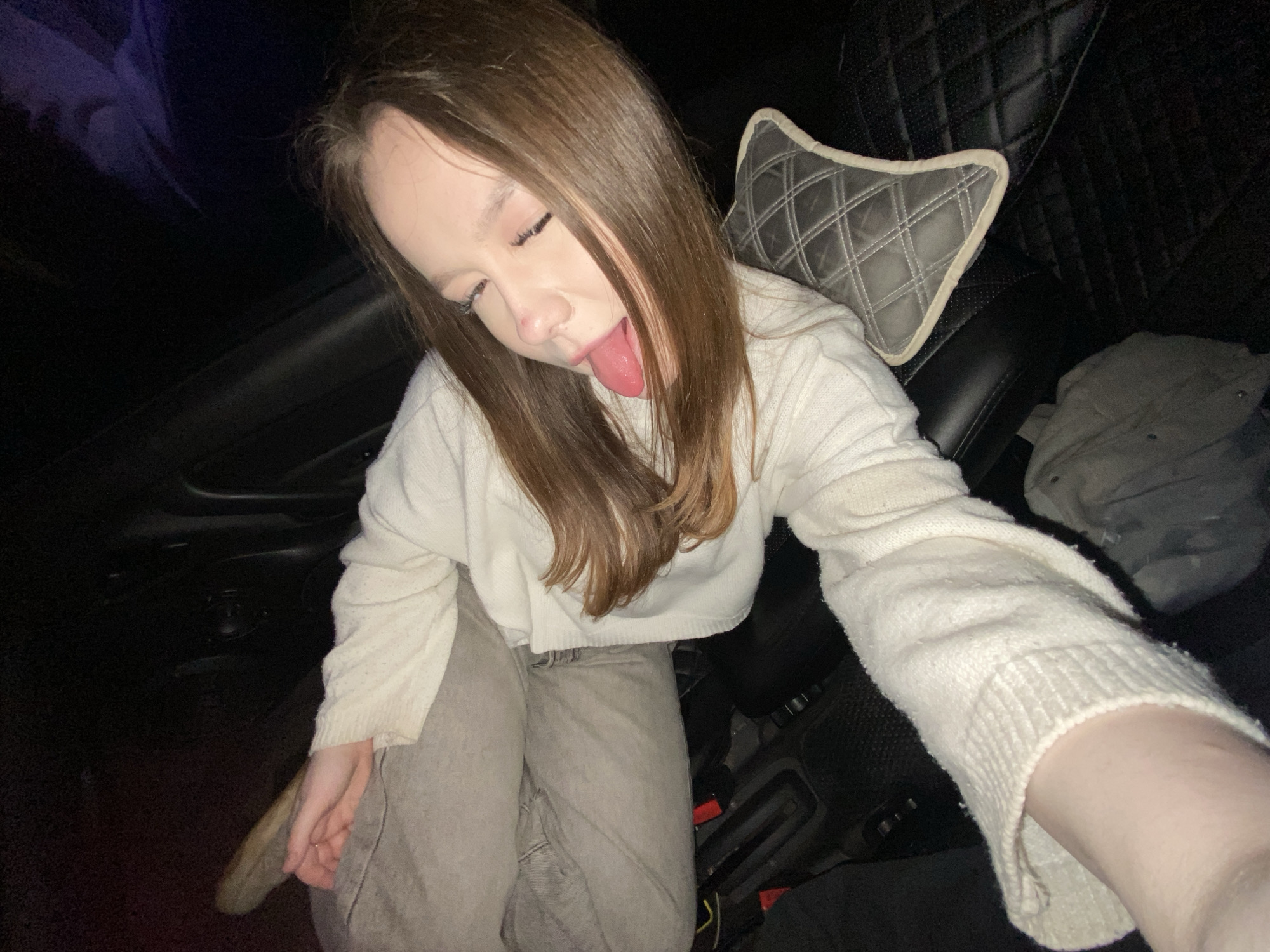 A young person, likely female, with light brown hair, is captured in a playful selfie inside a car at night or in a dimly lit environment. She is wearing a cream or off-white knitted sweater and light gray pants, leaning back in the car's dark, possibly leather, seats. Her eyes are closed or squinting, and she is sticking out her tongue, conveying a lighthearted and silly mood. Her left arm is extended towards the camera, indicating she is taking the photograph herself.
The car's interior features a dark background, with details such as a gray, diamond-patterned cushion with light trim placed on the seat. The car seats themselves also appear to have a quilted pattern. Red seatbelt buckles are visible near the floor of the vehicle. A dark gray object, possibly clothing, is next to the cushion. The general setting suggests a casual moment, possibly while traveling or parked in Chaikovsky, Russia. No visible text is present in the image.