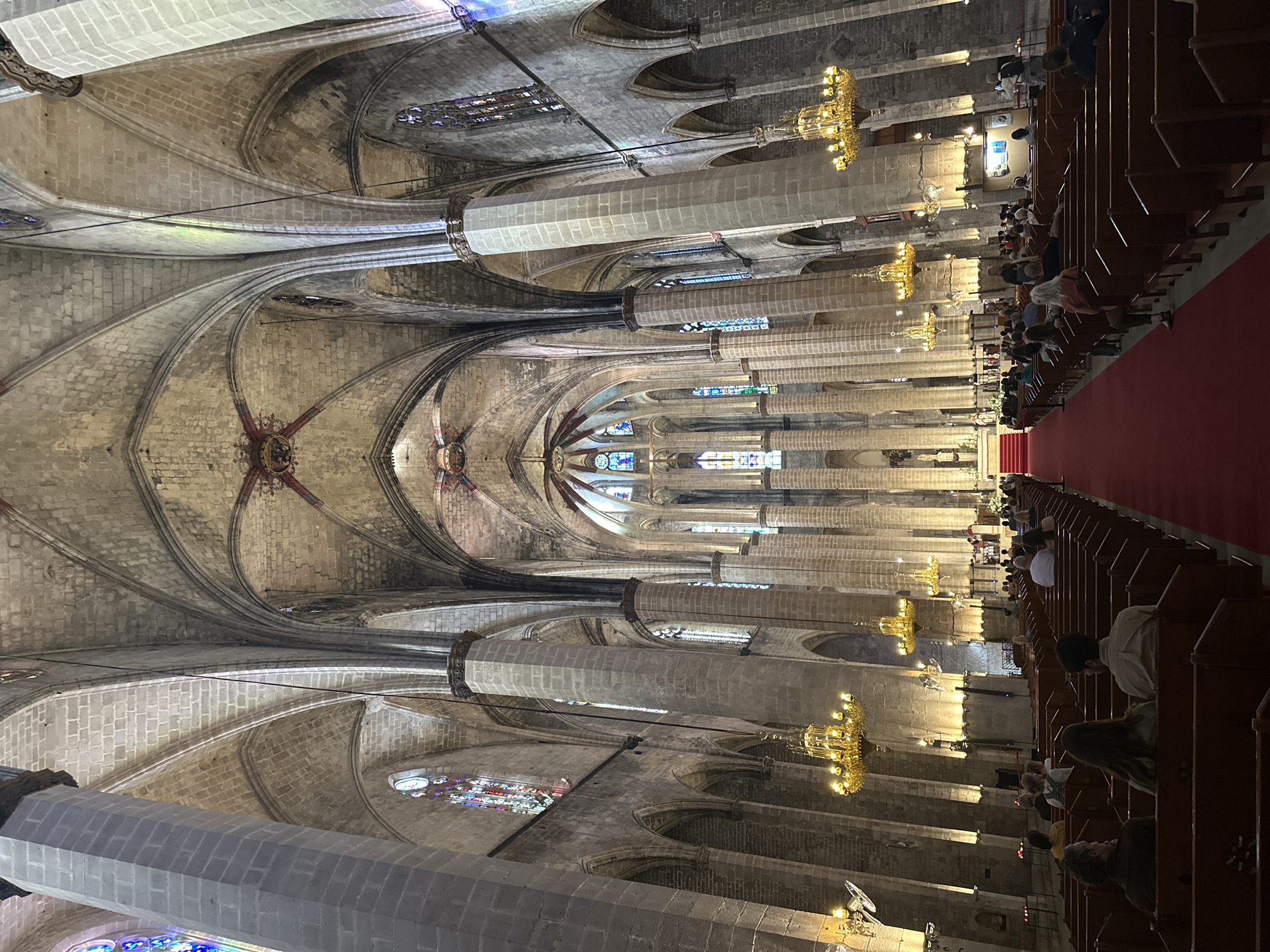 The image depicts the interior of the Basilica of Santa Maria del Mar, located in Barcelona, Catalonia, Spain. The view is from the rear of the central nave, looking towards the main altar at the eastern end. The structure features extremely tall, slender octagonal stone columns supporting a high, grey stone ribbed vault ceiling. Numerous pointed arches delineate the nave and side aisles. Stained-glass windows, some circular and others rectangular, are visible high on the upper walls, admitting colored light. The stone walls exhibit a textured, aged appearance.
A wide, red carpet extends down the central aisle, flanked by rows of dark wooden pews on both sides. Approximately 50 to 70 individuals, predominantly adults, are seated in these pews, facing the altar. Their posture suggests quiet observation or participation. In the foreground, several individuals are seen from behind or the side. The altar area at the far end is elevated by several red-carpeted steps, with a light-colored altar structure. A small group of people is visible near the altar, with one individual standing at a podium.
Illumination sources include natural light through the stained glass, casting colored reflections, and multiple ornate golden chandeliers suspended from the ceiling. Additional artificial uplighting fixtures mounted on the lower side walls cast warm light onto the stone. A thin black cable descends vertically from the ceiling in the mid-ground. The overall environment is a large, historic Gothic church interior during what appears to be a service, event, or quiet visiting period.