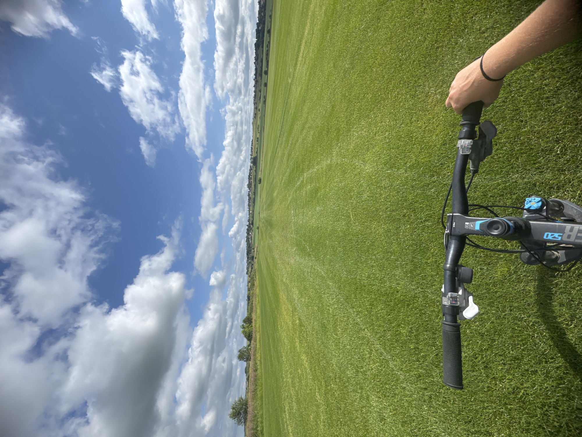 The image is a first-person perspective from a bicycle, showing a wide expanse of neatly mown green grass, likely a park or a sports field. The field stretches towards a distant treeline and a cluster of buildings on a gentle incline under a bright blue sky filled with scattered white clouds. The handlebars of the bicycle are visible in the foreground, with a hand holding the right grip. The bicycle itself appears to be a mountain bike, with visible components like gears and brakes. The overall scene suggests an outdoor recreational activity on a sunny day. The image does not provide enough information to place it in Kempelevo, Russia.