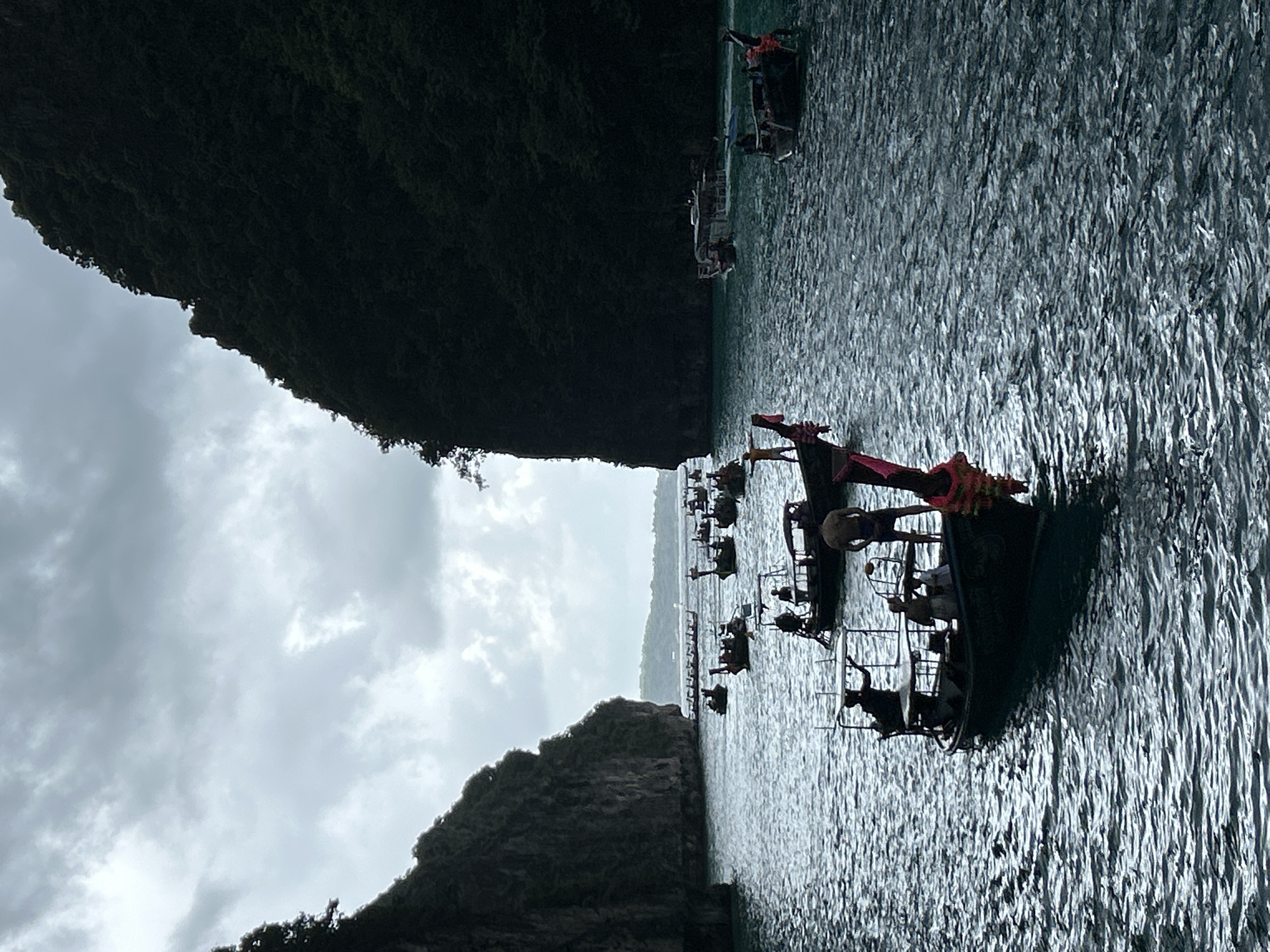 The image shows a body of water with several longtail boats. Two large, dark, tree-covered cliffs are on either side of the water, creating a narrow passage. The sky is overcast with dramatic clouds. In the foreground, a longtail boat carries several silhouetted figures. Further in the distance, more longtail boats are visible, some with passengers. The water is choppy and reflects the dim light. The scene appears to be a scenic bay or cove, possibly in the area of Haad Yao, Thailand, which is known for its limestone cliffs and boat tours.