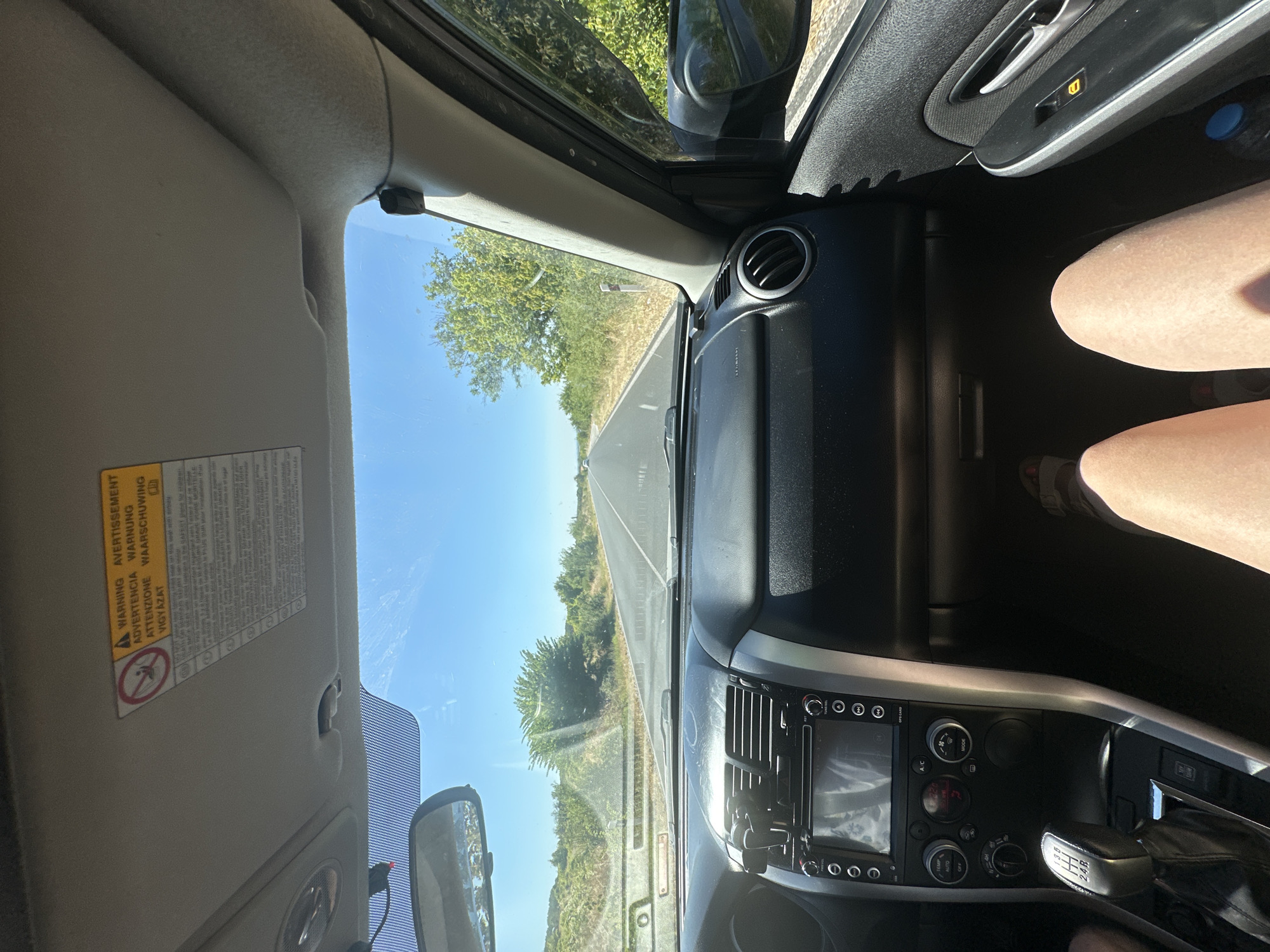 The image shows the interior of a car looking forward through the windshield. The dashboard is visible, with a GPS unit, air vents, and gear shift. Outside, a paved road stretches ahead, flanked by trees and vegetation under a clear blue sky. The scene is captured from the passenger seat. The image does not contain any information that allows for the identification of the location as Tatarevo, Bulgaria.