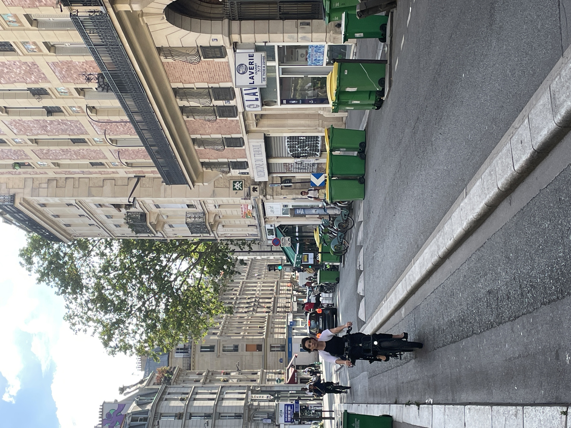 The image captures a vibrant daytime street scene in Paris, France. The perspective is slightly elevated, looking down a city street flanked by multi-story buildings of classic Parisian architecture, characterized by light-colored stone facades, numerous windows, and decorative iron balconies. Lush green trees line the left side of the street, partially obscuring the buildings and framing a bright, partly cloudy sky.
In the foreground, a person is riding a black bicycle directly towards the viewer, seemingly smiling or acknowledging the camera. They are wearing a white top with dark sleeves or a dark vest. Further down the street, other individuals on bicycles and pedestrians can be seen, along with a parked black car. The street itself is paved asphalt, with a clear curb and sidewalk running along the right side. A row of large green garbage bins with yellow and green lids is neatly lined up along the sidewalk on the right, extending into the background.
Visible signs on the buildings include "LAVERIE" (laundry), which specifies "OUVERT 7/7 DE 7H30 A 22H00" (Open 7/7 from 7:30 AM to 10:00 PM). Another sign reads "MEDECINE DU TRAVAIL" (Occupational Medicine). Other partial texts like "Lycas," "RESSING" (likely part of Pressing, dry cleaning), and "CAR SAR" are also discernible. A blue directional sign with a white arrow is visible further back. The overall impression is that of a busy, yet pleasant, urban environment during clear weather.