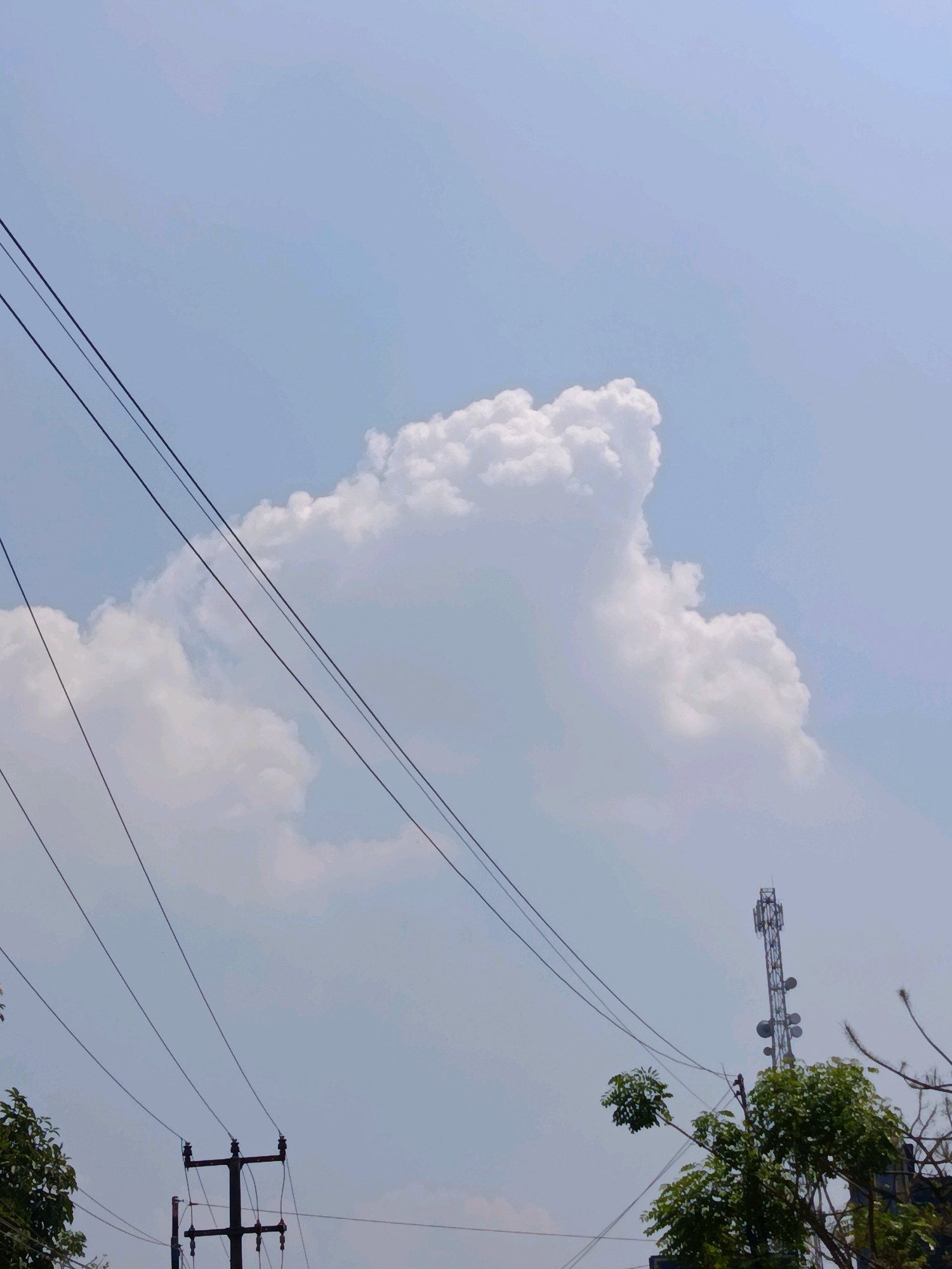 The image depicts a sky with scattered clouds, a prominent cumulus cloud formation centrally located, and several power lines stretching diagonally across the frame. In the lower right, a cellular tower is visible, surrounded by trees. The scene is set in Babelan, Indonesia, and is captured on a clear day. No people are present in the image.