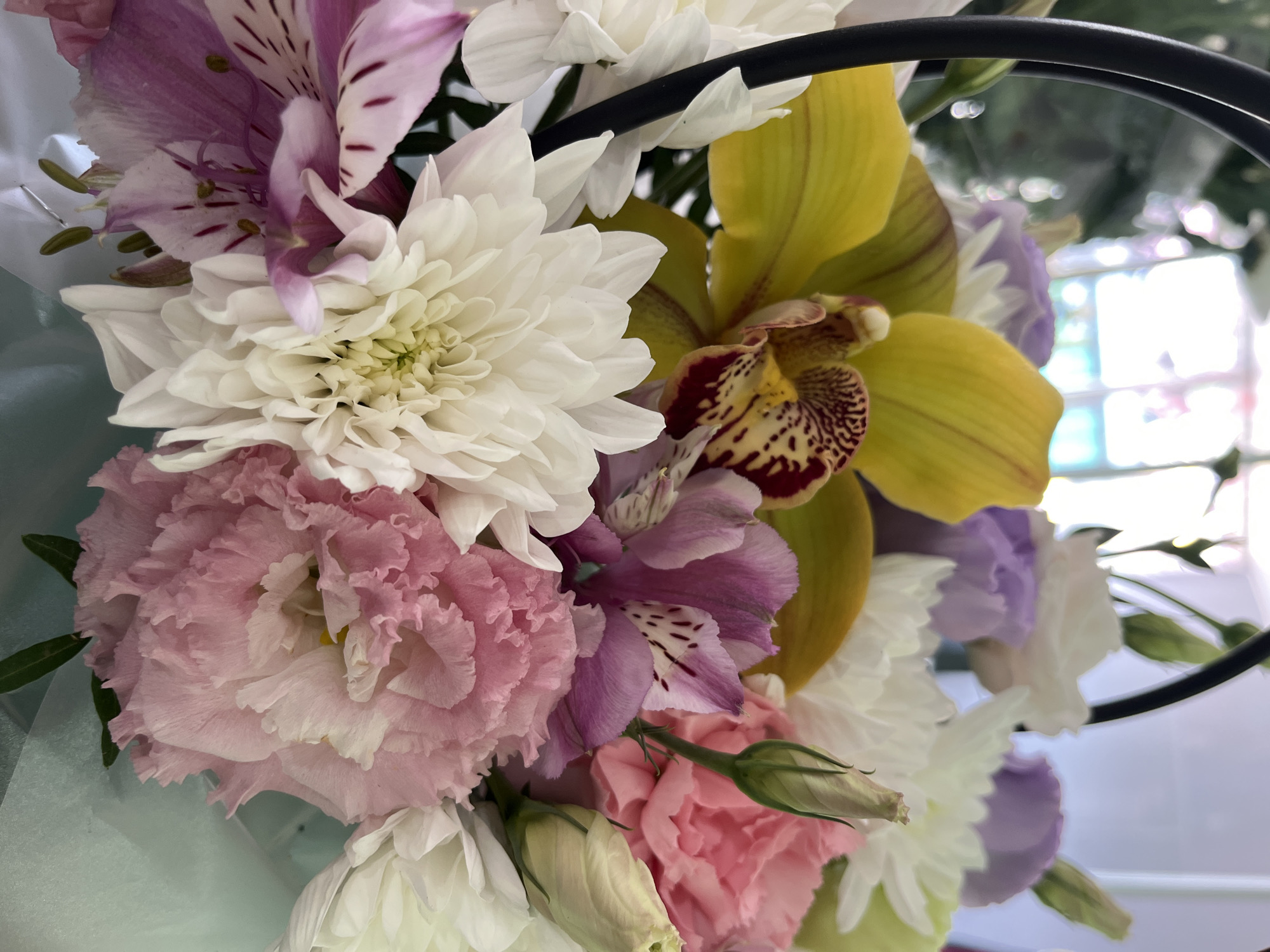 A close-up, eye-level shot captures a vibrant floral arrangement. The bouquet is predominantly composed of a large, bright yellow orchid with deep burgundy and yellow speckled markings. Surrounding the orchid are several white chrysanthemum-like flowers, characterized by their numerous delicate petals. Interspersed among these are soft pink carnation-like flowers with ruffled edges and a few lighter purple lisianthus flowers. The background is softly blurred, showing hints of light and possibly a window or other room interior. The overall impression is one of a colorful and delicate floral display.