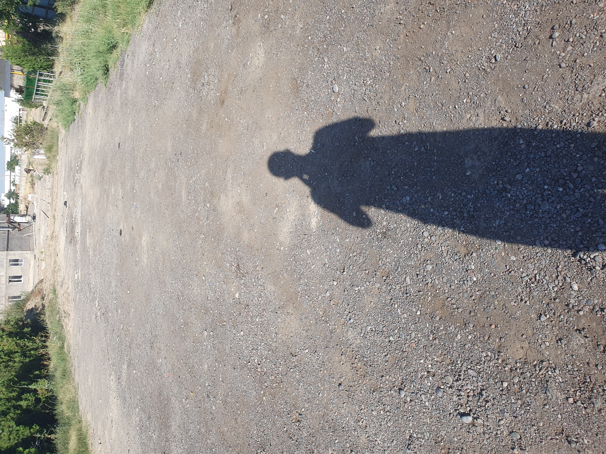 A ground-level, full-body shot of a person's shadow is cast on a rough, gravelly surface. The shadow is long and distinct, indicating a strong light source from the front and to the side. The person appears to be standing, with a suggestion of arms and possibly holding something. The environment is an outdoor, unpaved area, likely a street or construction site, with scattered stones and patches of dirt. In the background, partially visible are buildings and vegetation, including trees and grass, which appear to be part of an urban or suburban landscape. The overall scene suggests a bright, sunny day. This image could have been taken in a location such as the construction site of the Tashkent City International Business Center, in Tashkent, Uzbekistan, which features extensive areas of unpaved ground, scattered building materials, and nearby urban development.