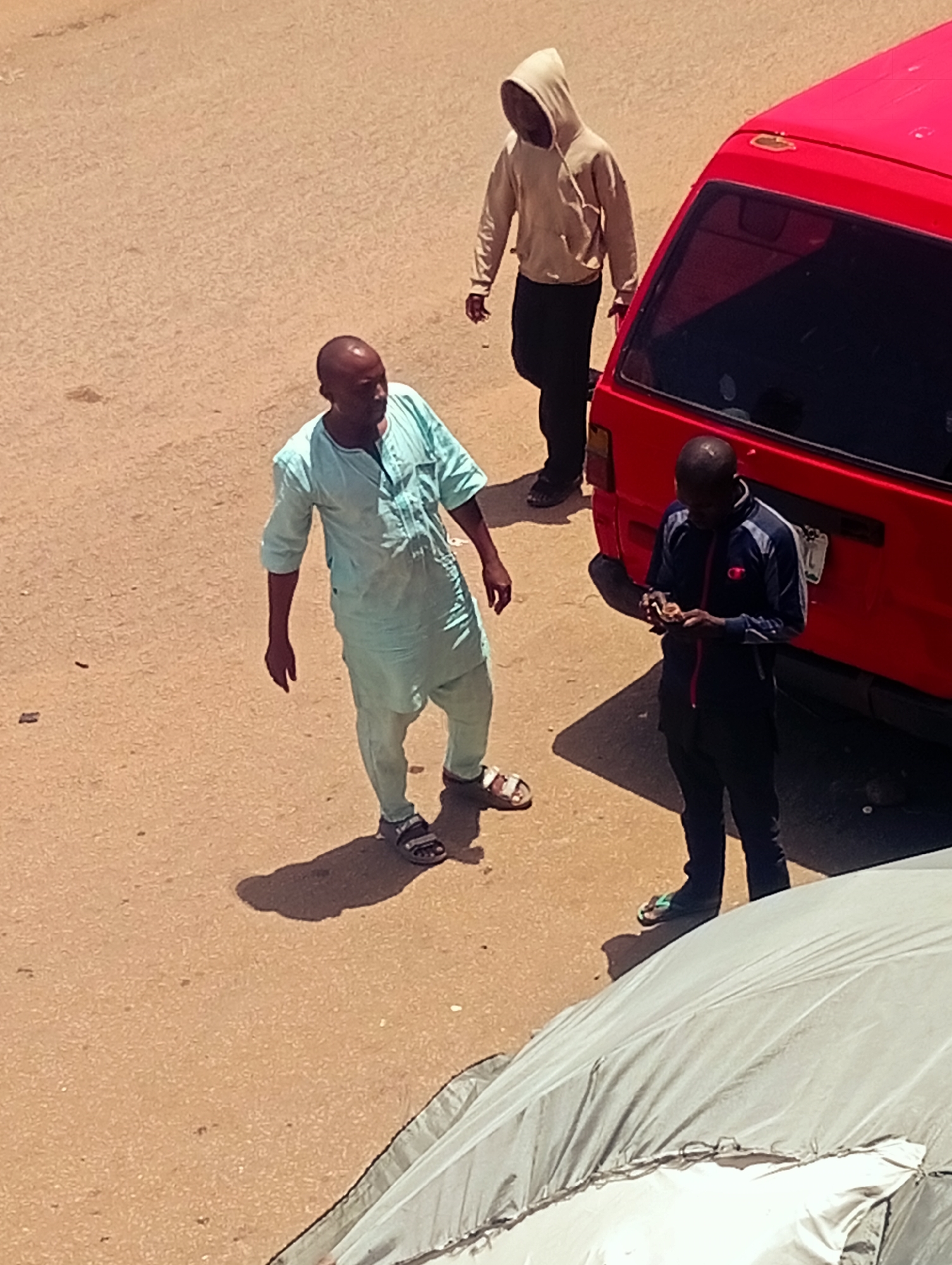 The image shows an outdoor scene with three individuals and a red van. The ground appears to be a paved surface.
One man is wearing a light blue traditional tunic and trousers, along with sandals. He stands facing forward with his arms slightly outstretched. A second man is wearing a hoodie and black trousers, walking away from the camera. The third man is wearing a dark jacket with a red zipper, dark trousers, and sandals. He is standing near the back of a red van. He appears to be looking at something in his hands. A partial view of a gray tarp is visible in the foreground.