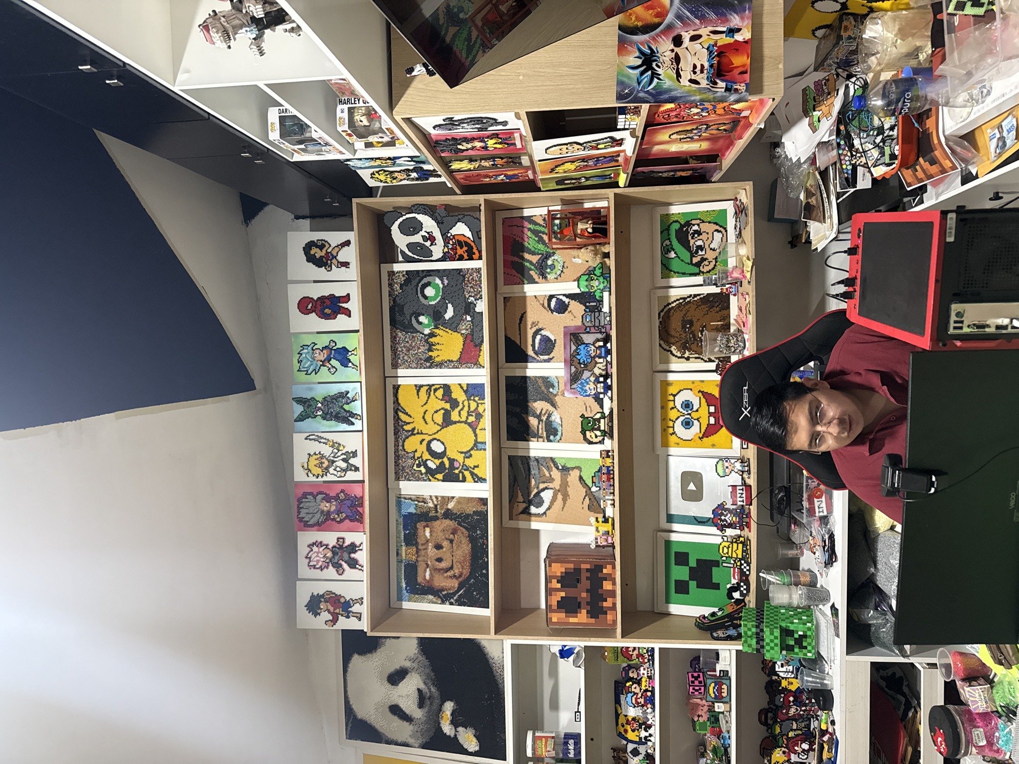 A medium shot depicts a person seated in front of a computer setup, presumably in Puebla, Mexico, given the prompt. The individual has dark hair and is wearing a red shirt. The computer setup includes a black monitor, a red and black PC tower, and a black gaming chair.
Behind the person, a wooden shelving unit is filled with pixel art pieces, some of which depict characters from popular culture like Pokémon, Dragon Ball Z, One Piece, and Minecraft. Other shelves display Funko Pop figures, manga, and various craft supplies. The walls are painted white and blue. The room is cluttered with various items, including loose beads and other crafting materials. The image has been rotated.