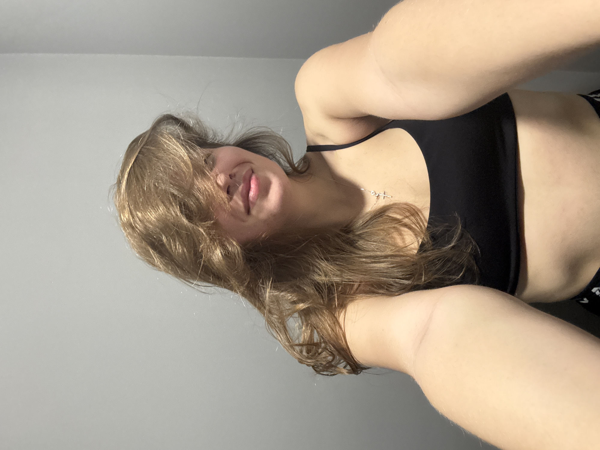 A young woman is seen from a low angle, likely a selfie. She has shoulder-length brown hair that partially obscures her face, and she is sticking her tongue out slightly. She is wearing a black sports bra and a silver cross necklace. The background is a plain gray wall. The location is not identifiable as Belgrade, Serbia based on the provided image.