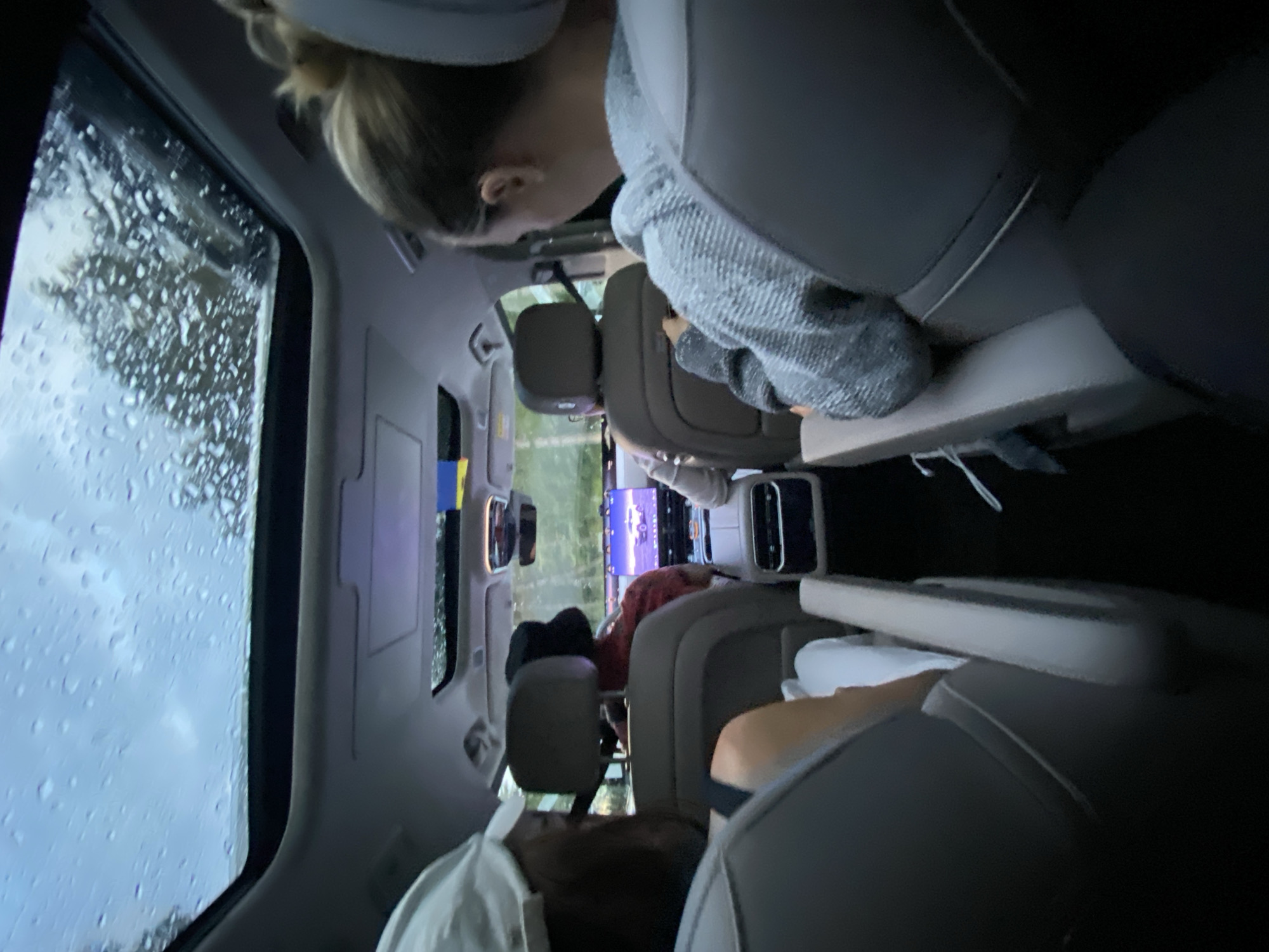 The image captures the interior of a vehicle from a rear-facing perspective. Four individuals are visible, seated in what appear to be second-row and third-row seats. The roof of the car features a sunroof with visible raindrops on the exterior, suggesting inclement weather. A central touchscreen display is prominent in the front of the car. The passengers are engaged in their own activities, with one individual in the foreground looking at a device and another in the middle row appearing to use a phone. The lighting suggests it is daytime, possibly overcast. There is no discernible information in the image that places the scene in the city of Ustia, Moldova.