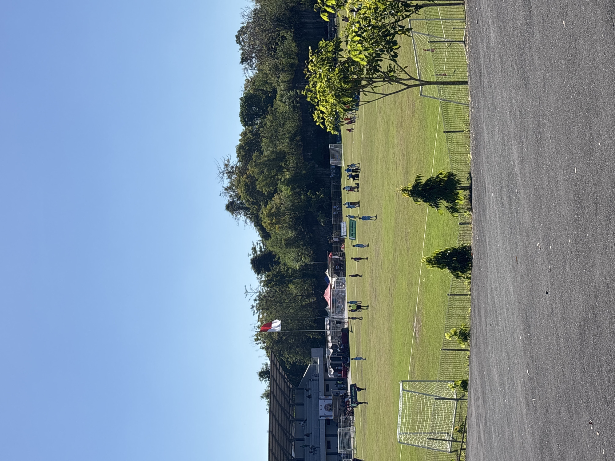 The image shows a soccer field in Kuta Selatan, Indonesia. The field is green, with white lines marking the boundaries. Two soccer goals are visible on either end of the field. A team of players wearing blue uniforms is practicing on the field. Spectators are in the bleachers next to the field. A red and white Indonesian flag is flying. The weather is sunny with a blue sky. The field is surrounded by trees and asphalt.