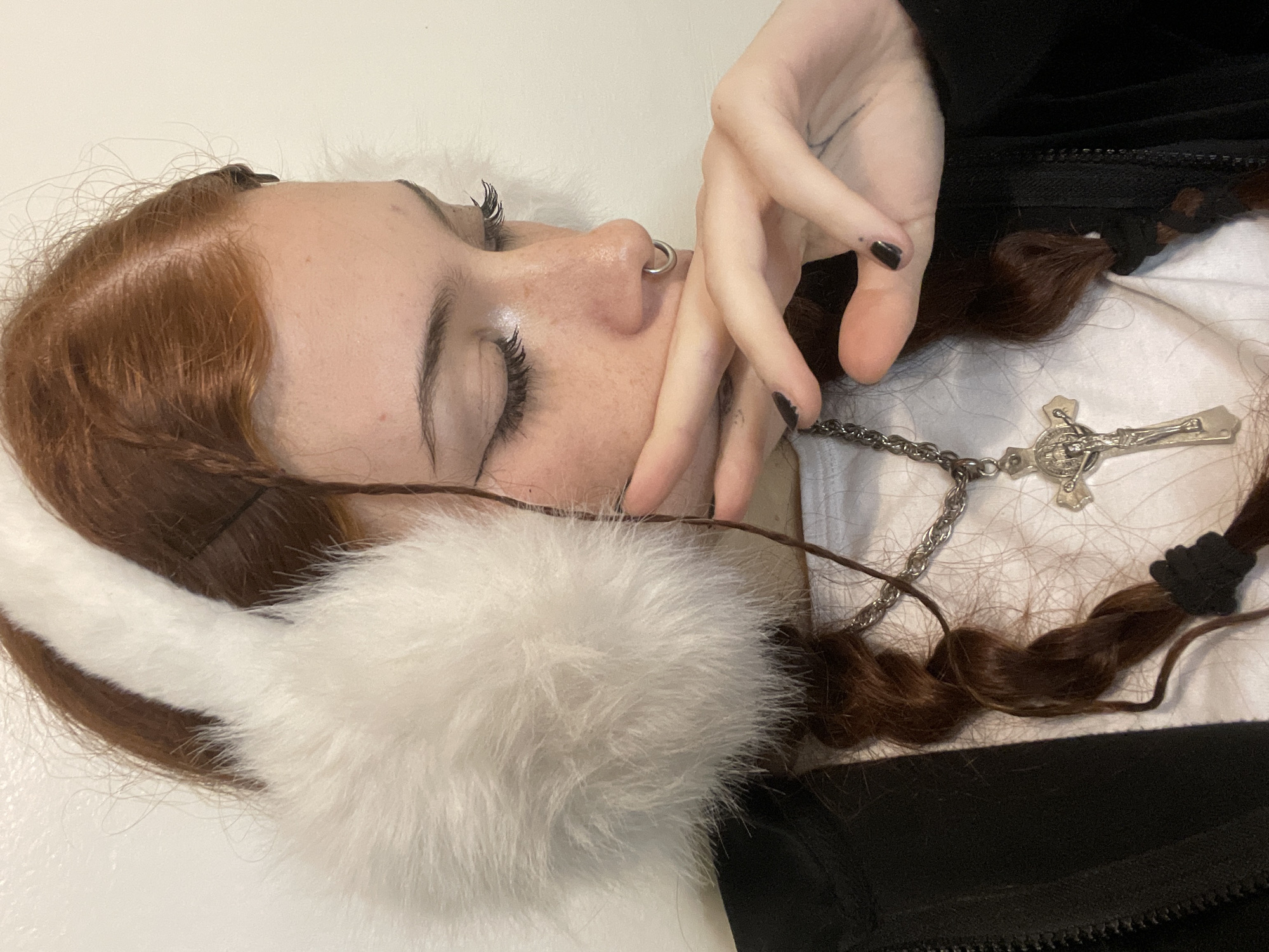 A young woman is shown from an aerial perspective, lying on her side on a white surface. She has red hair styled in braids. She is wearing white fluffy earmuffs and a silver chain necklace with a cross pendant. Her eyes are closed, and her hand is near her face. She has a nose ring, and her fingernails are painted black. She is wearing a white shirt with a black jacket on top. The image provides no information about the city of Buenos Aires, Argentina.