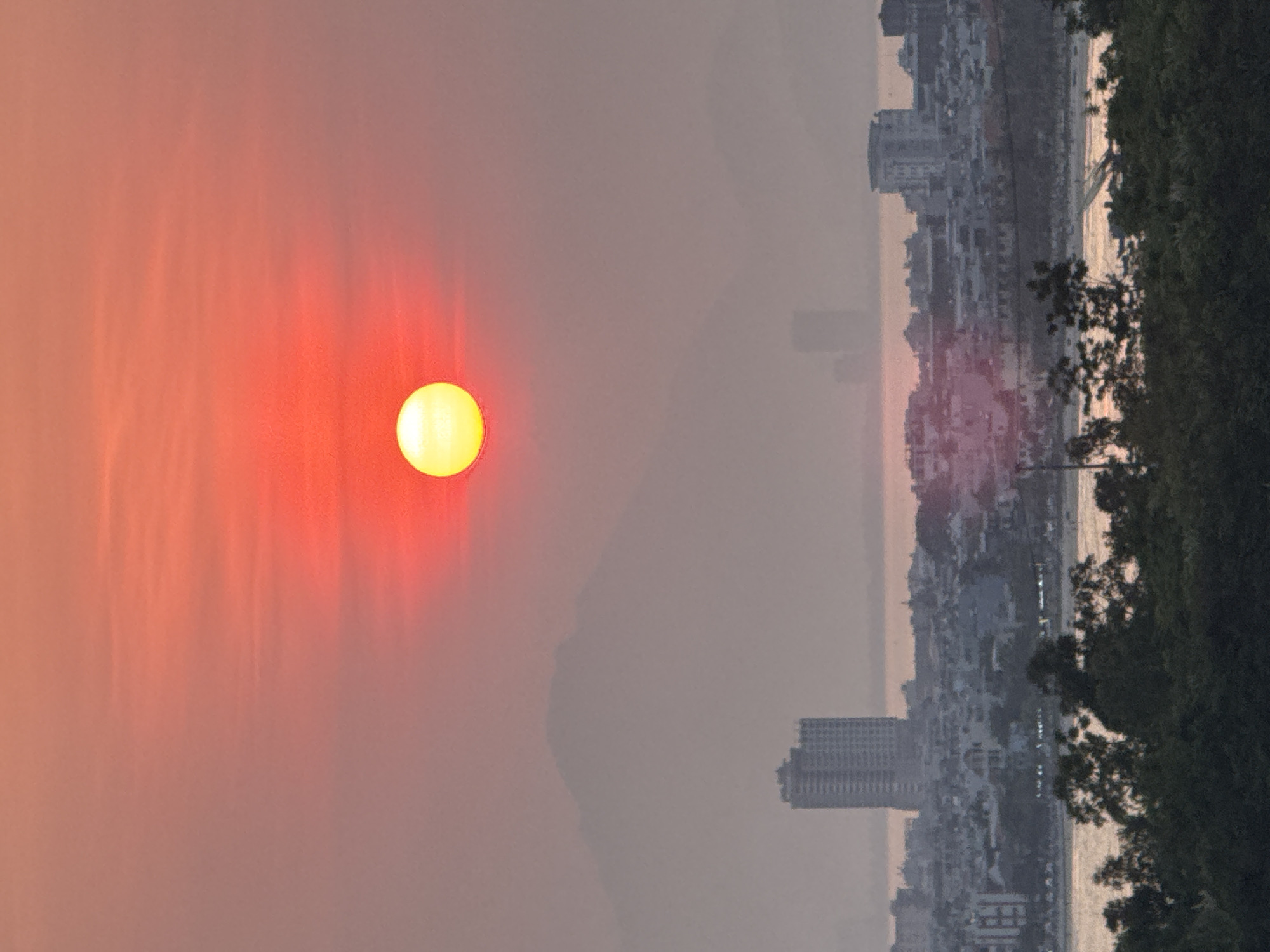 The image depicts a hazy sunset over the city of Đà Nẵng, Vietnam. In the sky, a large, orange-red sun is visible. Below the sun, a silhouette of distant mountains can be seen. The city skyline is obscured by haze, but buildings and structures can be identified. A body of water borders the city. A dark mass of trees is in the right foreground.