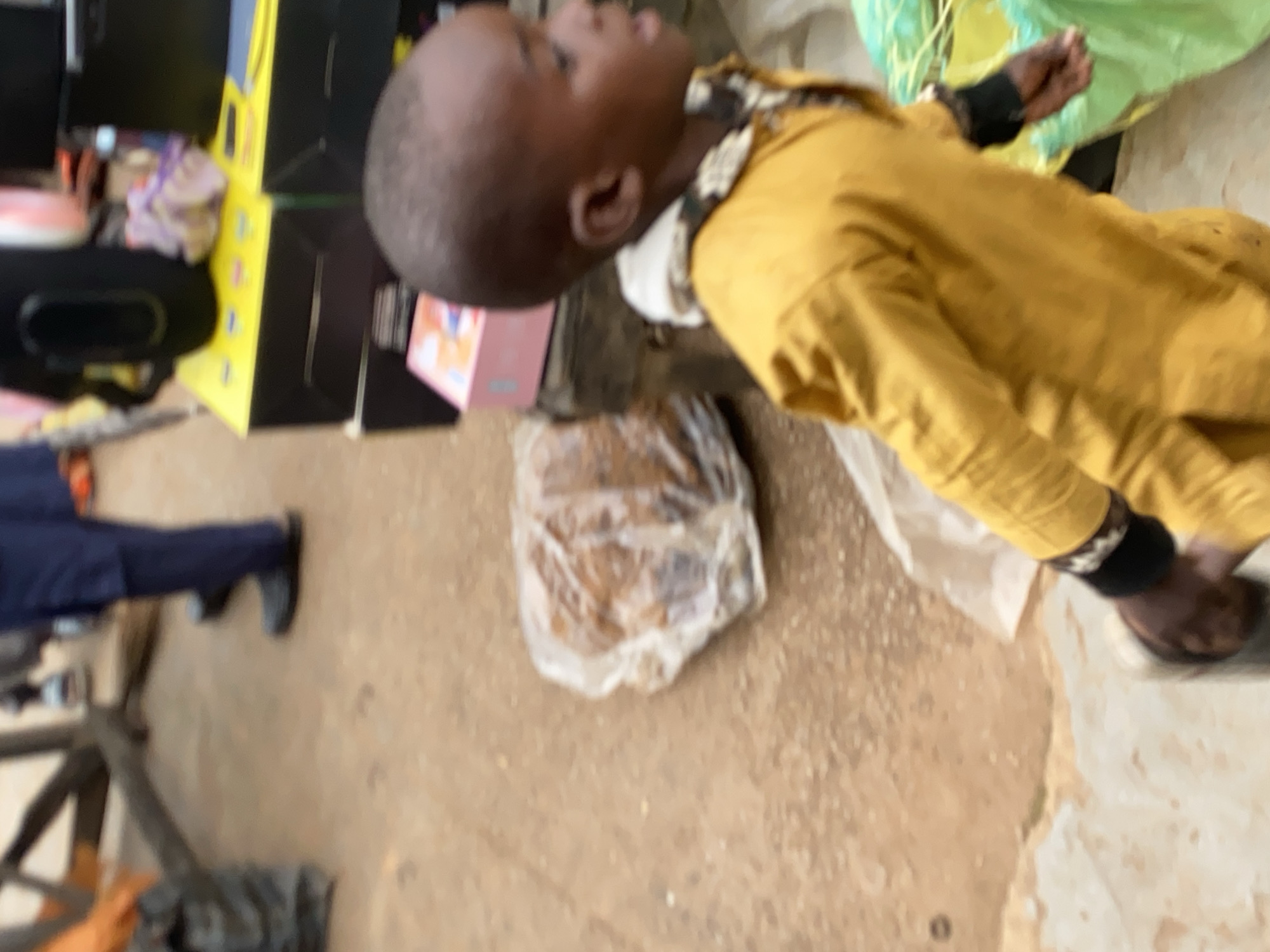 A child in a yellow garment stands in a marketplace. The child is looking to the right of the frame. In the background, there are yellow and black boxes stacked. To the left of the child, there is a plastic-wrapped item. The ground is dirt. The scene appears to be in Bukuru, Nigeria.