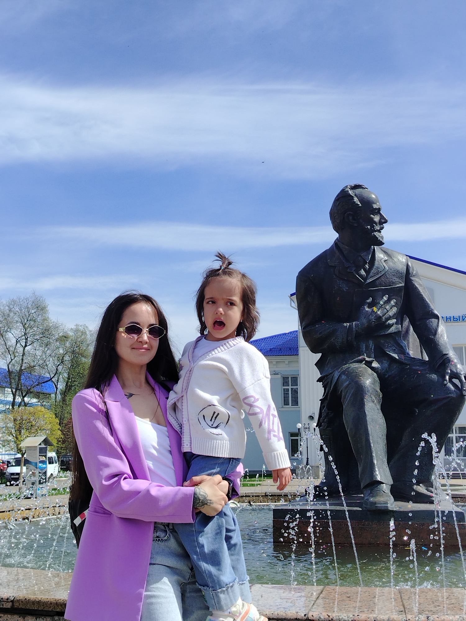 The image captures a woman holding a young child in front of a fountain in Chaikovsky, Russia. A bronze statue of Anton Chekhov sits in the background behind the fountain. The woman wears a purple blazer, jeans, and sunglasses. The child is dressed in a white sweater and jeans. The sky is partly cloudy and the surrounding area features trees and buildings.