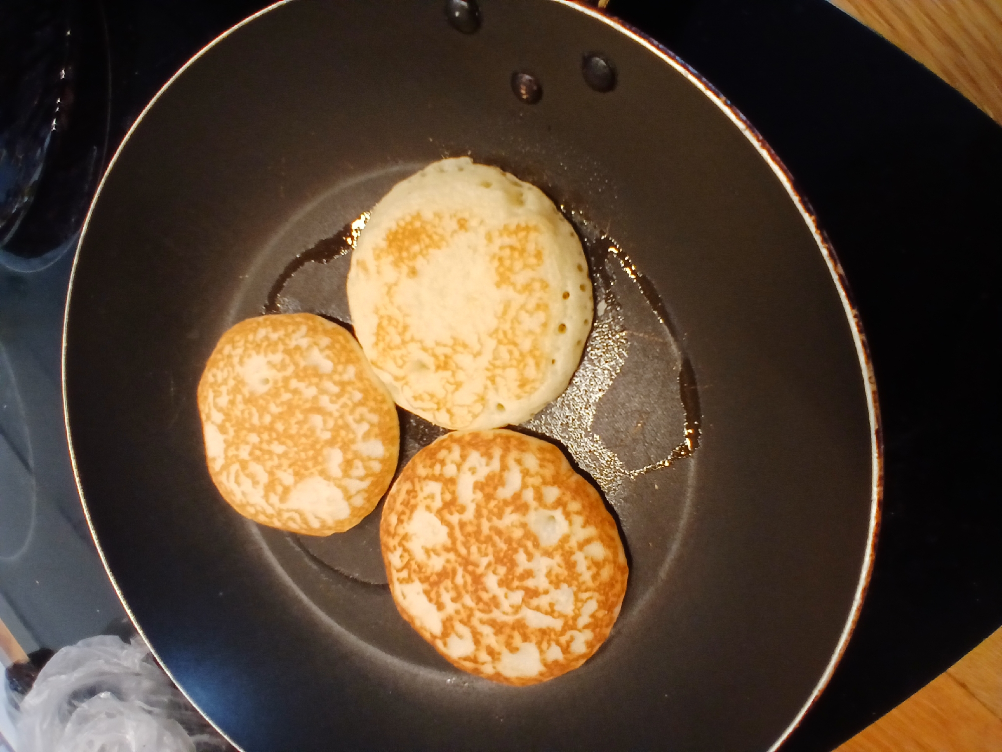 Three golden-brown pancakes are cooking in a black non-stick frying pan on a dark stovetop. The pancakes are circular and have a slightly uneven surface texture. The pancake on the left is a richer golden brown with a more pronounced texture, while the other two are a lighter golden color and appear softer. There is a small amount of glistening oil in the pan around the pancakes. The pan is round with a black handle. In the upper left corner of the image, a crumpled white plastic bag is partially visible. The background shows a dark, reflective surface, possibly part of the kitchen counter or appliances. The lighting suggests an indoor setting, likely a kitchen. The scene does not provide any specific details to determine its location in the city of Yekaterinburg, Russia.