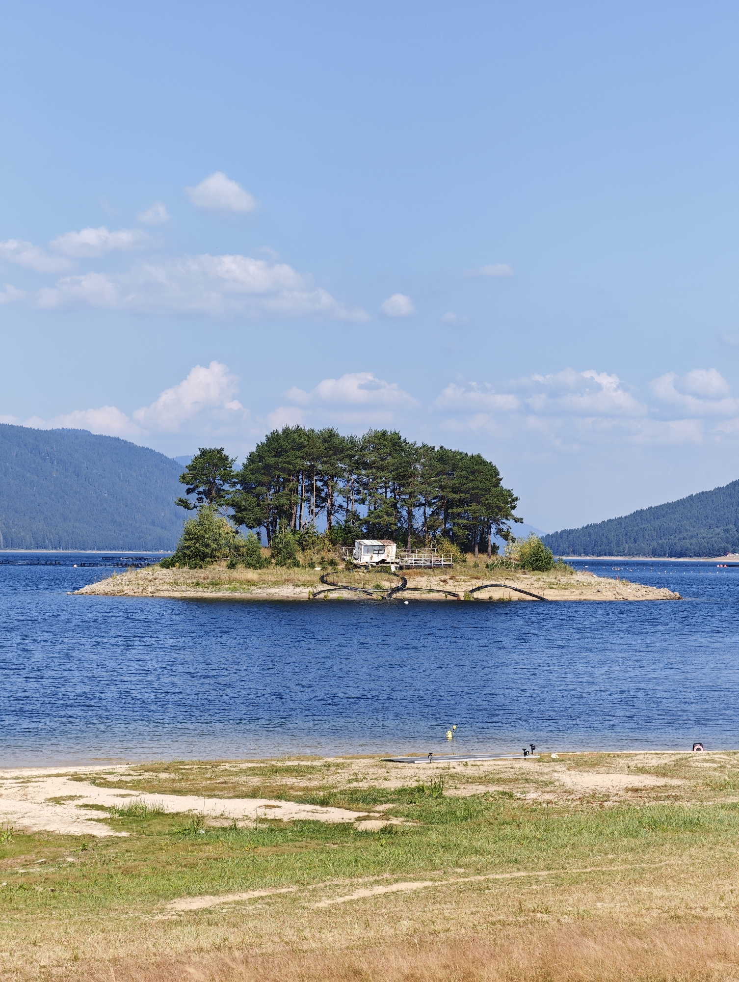 The image depicts a picturesque scene of a small, tree-covered island in the center of a large, blue body of water, likely a reservoir. In the foreground, a sandy beach transitions into a grassy area with dry vegetation. The island itself is densely populated with tall, green coniferous trees, creating a natural canopy. A small, white structure, possibly a caravan or a small building, is visible on the island, along with some wooden railings and what appears to be a pathway leading to it. The water is calm with gentle ripples. In the background, rolling hills covered in dense forests rise up, their slopes meeting the clear blue sky dotted with fluffy white clouds. The overall impression is one of tranquility and natural beauty. The location is identified as the Dospat Reservoir in Dospat, Bulgaria.
