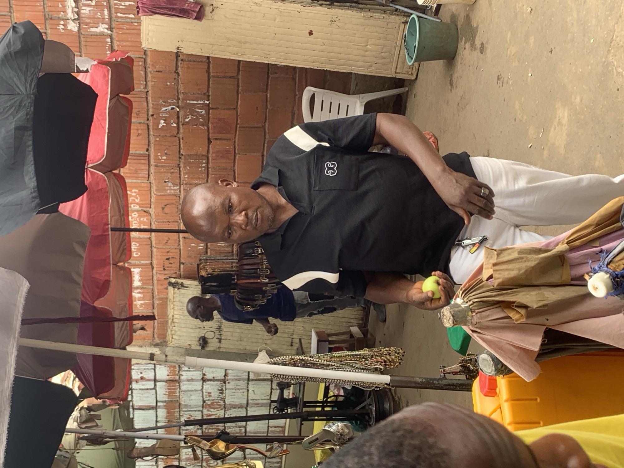 In an outdoor market setting in Abuja, Nigeria, a bald man with dark skin is holding a green apple in his right hand. He is wearing a black polo shirt with a white stripe on each shoulder and white pants. His left hand rests on his pants. Behind him, other vendors and market stalls are visible. A sewing machine is placed on a table to the left, and an assortment of colorful beaded necklaces hangs from a stand. A man in a blue shirt stands in the background to the left. The environment includes brick walls and several large umbrellas providing shade. A white plastic chair is situated to the right of the main subject, and a green plastic bucket is on the ground.