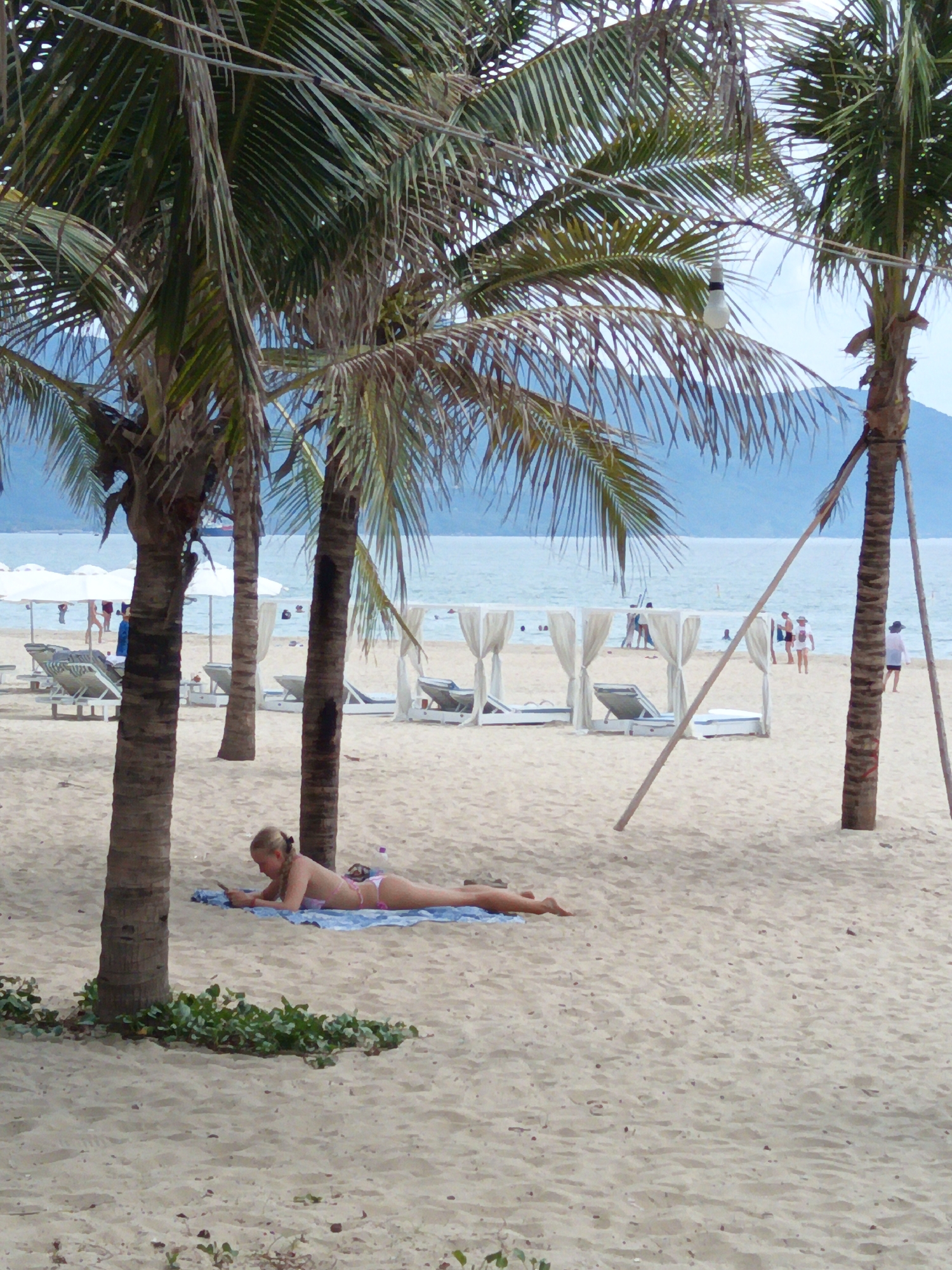 A blonde woman in a pink bikini lies prone on a blue towel on a sandy beach. She is looking at a phone. Three palm trees are in the foreground, two of which are tall and slender, and one is shorter and thicker. In the middle ground, there are white cabanas with white curtains, lounge chairs, and umbrellas. The ocean is visible in the background, with mountains in the distance. The image appears to have been taken in Da Nang, Vietnam.