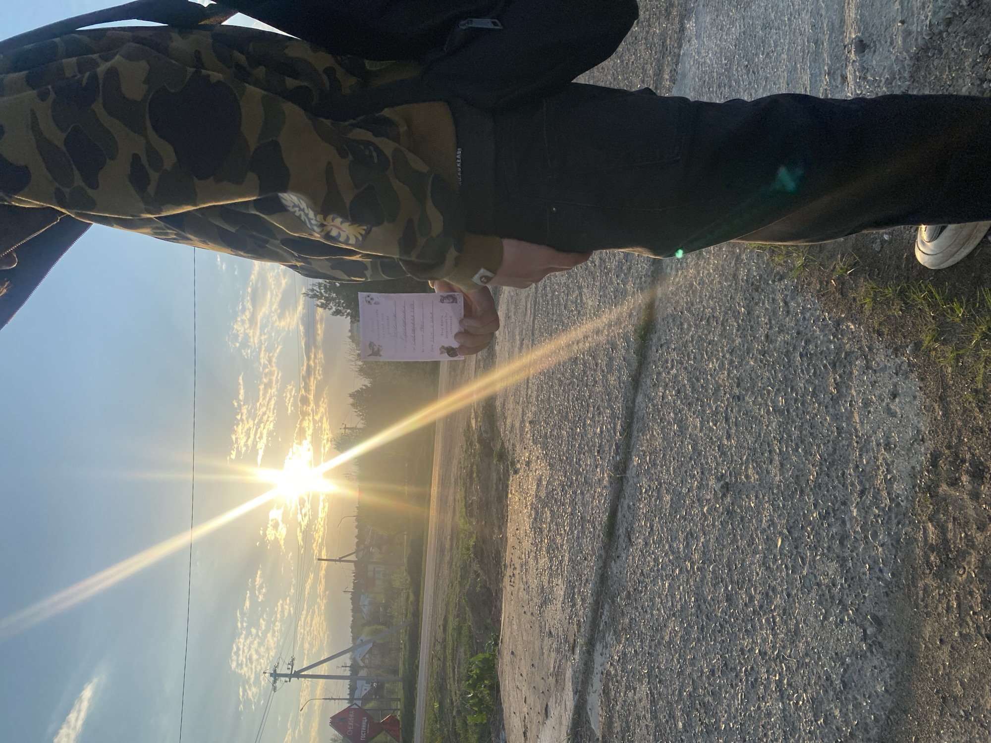A photograph taken in an area identifiable by a sign with the Cyrillic words "СНЕЖИНКА ГОСТИНИЦА" (Snowflake Hotel). The image focuses on a person standing on a road holding a card. The subject is wearing a camouflage patterned top, black pants, and a backpack. The photograph is taken outdoors, with the sun setting in the background, casting a bright glow. The setting includes a road, some greenery, and distant buildings, indicating a suburban or rural area.