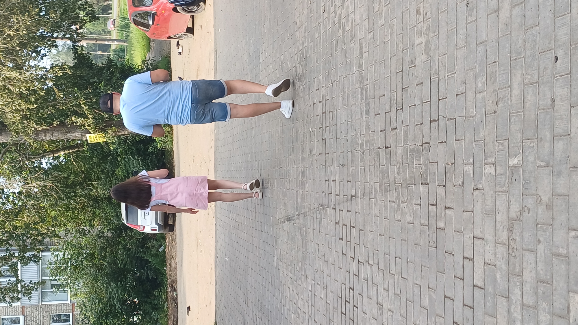 The image shows two people, a young woman and an adult male, walking away from the camera on a paved walkway. The woman is wearing a light pink pinafore dress over a white t-shirt and white sandals. The man is wearing a light blue t-shirt, denim shorts, and white sneakers. They appear to be walking in a city environment. In the background, there is a multi-story apartment building, trees, and a white SUV. To the right, a red car is partially visible. The scene is lit by daylight, suggesting it is daytime. The paved walkway is made of light grey interlocking bricks. There is a small bird on the ground in the middle left of the image. The location is identified as Izhevsk, Russia.