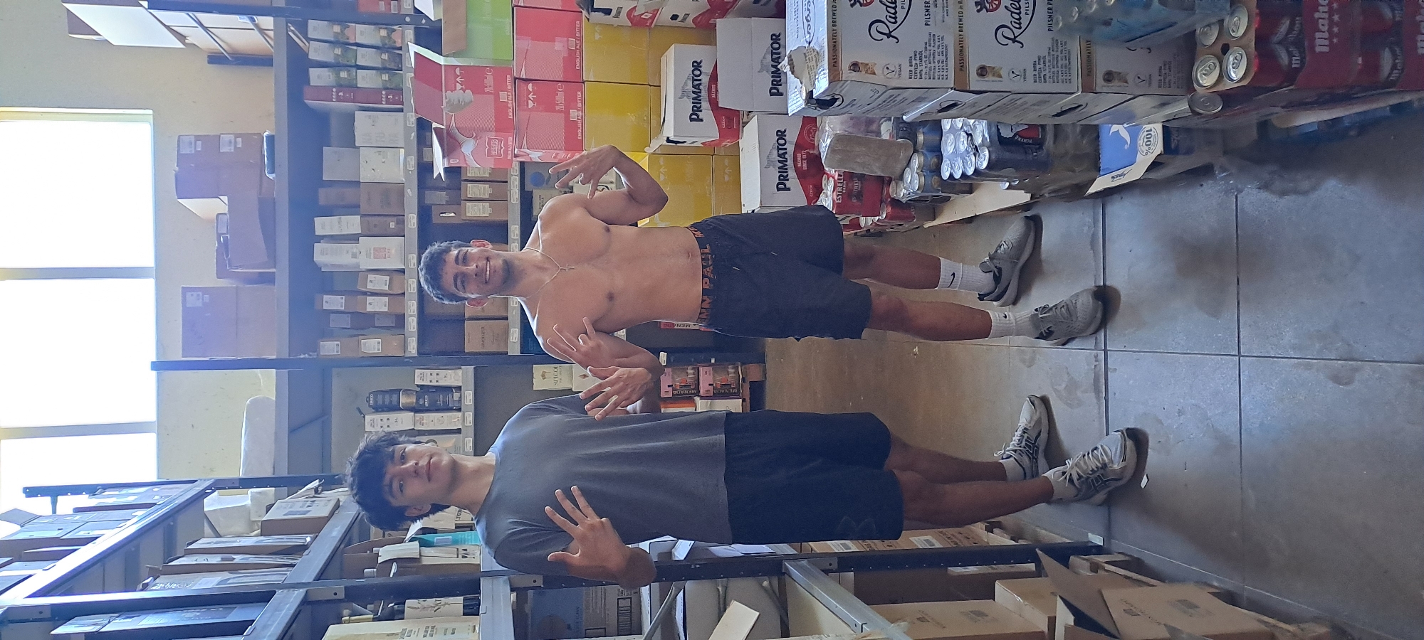 Two young men are standing in a warehouse. The man on the left is wearing a grey t-shirt and black shorts, and he is holding up his left hand, showing four fingers. The man on the right is shirtless, wearing black shorts, and holding up his right hand with four fingers extended. They are standing in an aisle between shelves stocked with boxes and bottles. The overall environment suggests a storage or distribution center, possibly for alcoholic beverages, given the branding on some of the boxes. The lighting appears to be natural, coming from windows high up on the wall.