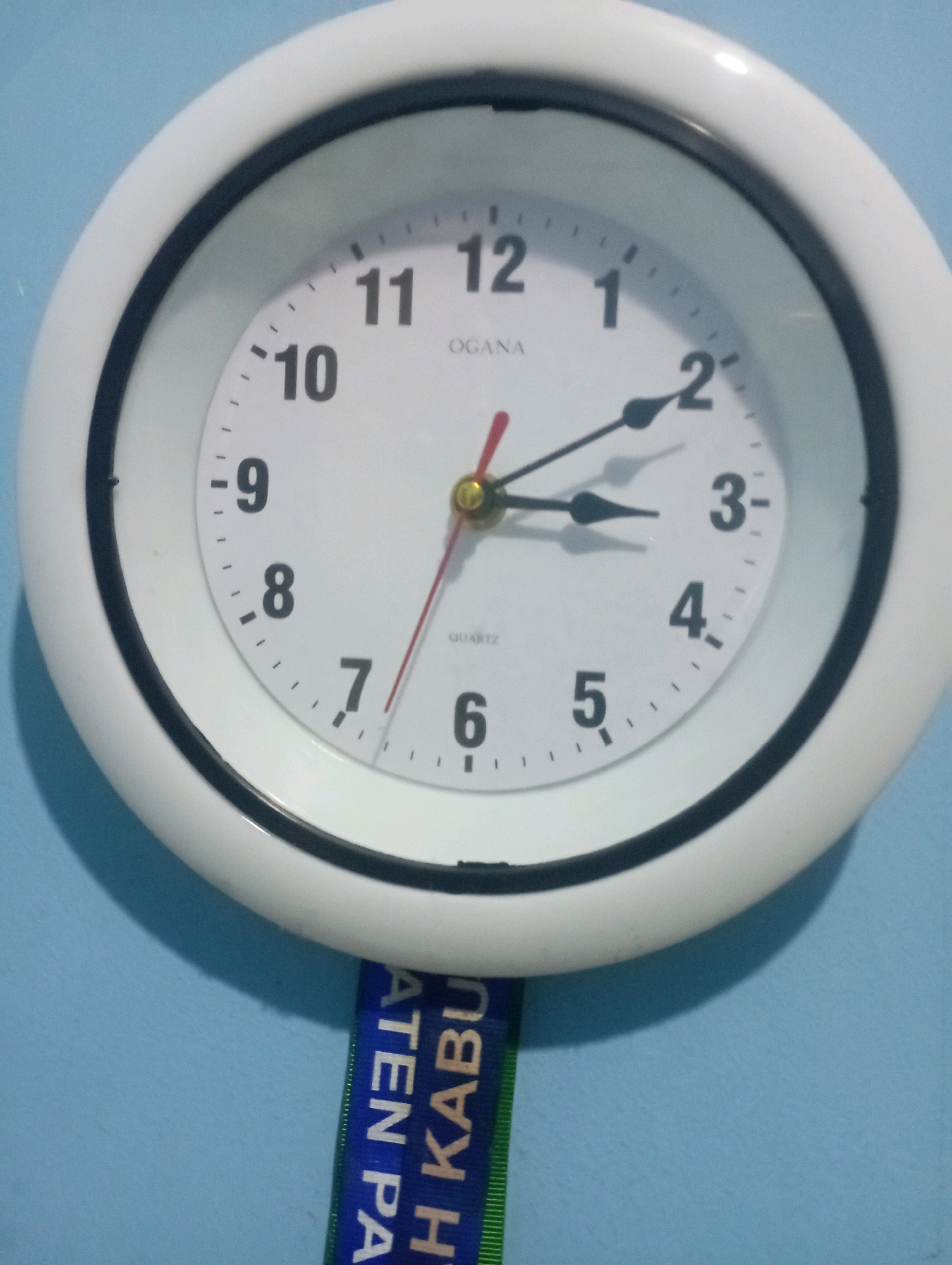 A round, white analog clock with a black bezel is hanging on a light blue wall. The clock face has black numbers and tick marks. The hour hand is pointing to the 2, and the minute hand is pointing between the 3 and the 4. A red second hand is also visible. Below the clock, a blue and green lanyard with white text is hanging down, partially obscuring the wall. The text on the lanyard appears to be in Indonesian. The scene is indoors, likely in a residential or office setting, given the commonality of the clock and lanyard. There are no people or animals present in the image. The lighting is consistent with indoor ambient light.