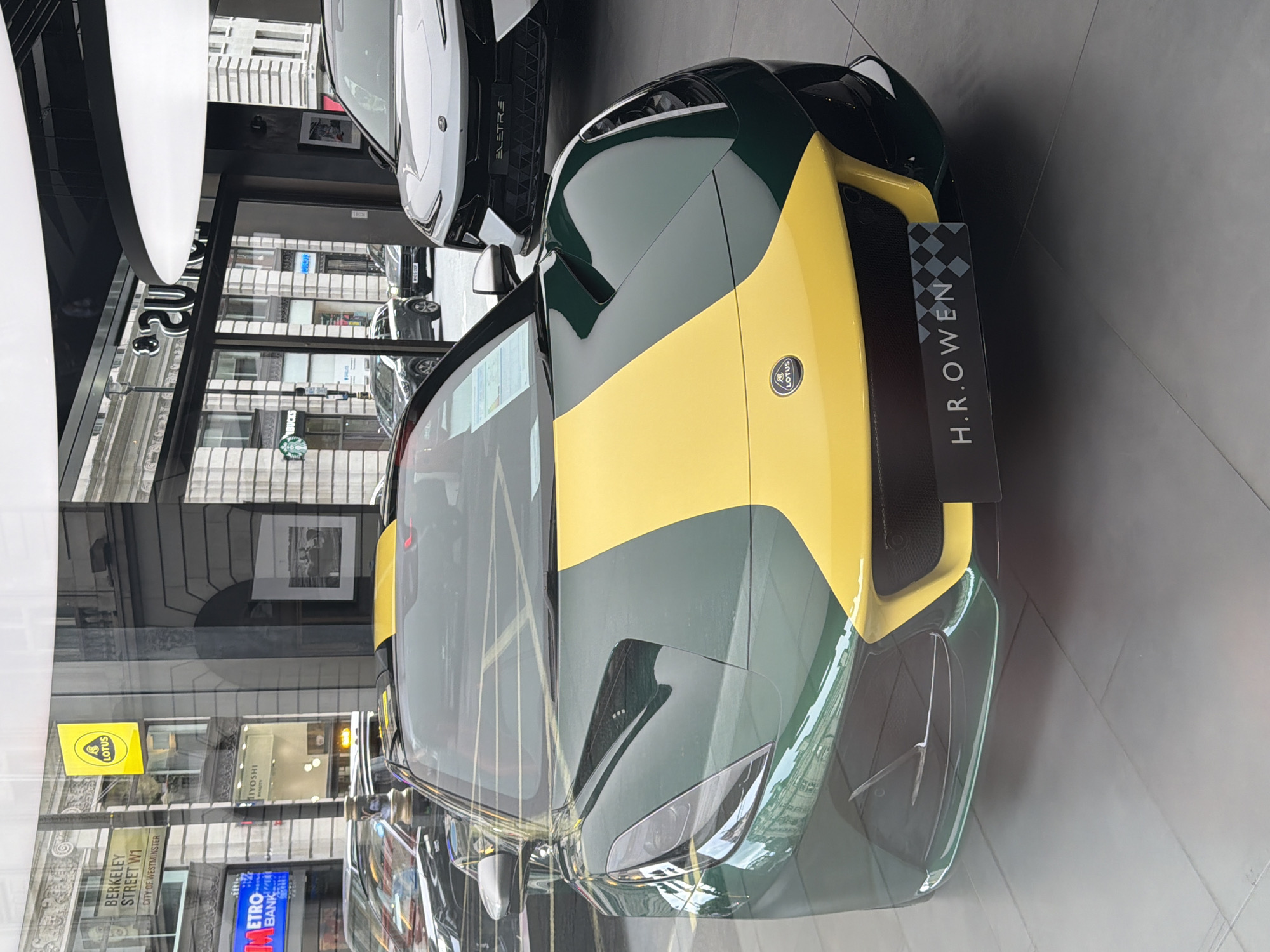 The image shows the front of a green and yellow Lotus sports car displayed in a showroom. The car has a prominent yellow stripe down the center of its hood. Behind the sports car, another white Lotus SUV is visible, with the word "ELETRE" visible on its front grille. The showroom appears to be located on Berkeley Street in the City of Westminster, London, United Kingdom, as indicated by signage visible through the window. The window also offers a view of a street scene with other cars and buildings, including a Starbucks. The showroom has a modern interior with a black ceiling and large pendant lights. The floor is tiled with large, light grey tiles. There are no people clearly visible in the image, though reflections in the glass suggest outdoor activity.