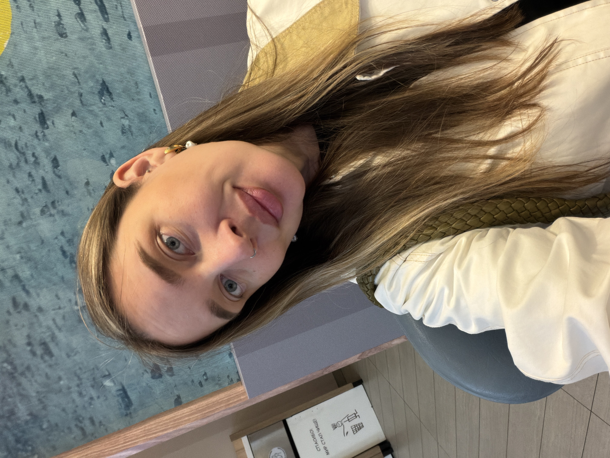 A close-up, slightly tilted portrait shows a young woman looking directly at the camera from an upward angle in an indoor setting. She has long, light brown hair with blonde highlights, light blue eyes, and well-groomed eyebrows. She wears a small silver nose ring and a gold-colored hoop earring with a white bead in her left ear. Her expression is calm with a hint of a gentle smile.
She is dressed in a white long-sleeved top or jacket, featuring a gold or khaki-colored lining visible around her collar and a textured, woven green detail on her right side. The background indicates a public indoor space, possibly a cafe or lounge. Elements visible include a gray upholstered chair, a wooden counter or shelf, and a patterned blue and grey floor or wall.
On the wooden surface, a white rectangular box is visible with Russian text that reads "СПАСИБО! МИР СТАЛ ЧИЩЕ!" (Thank you! The world has become cleaner!), beneath an icon of a person putting an item into a waste bin. The overall lighting is soft, suggesting an indoor daytime environment in Moscow, Russia.