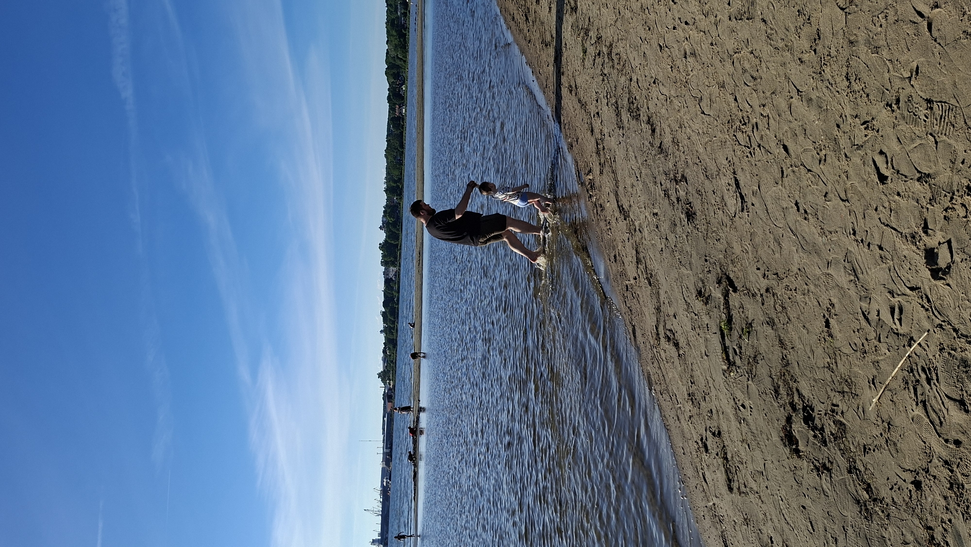 A man and a young child are walking in shallow water near a sandy beach under a bright blue sky with wispy clouds. The man, wearing a black t-shirt and dark shorts, is holding the child's hand. The child is wearing blue shorts. The water is calm with small ripples. In the background, several other people and dogs are visible on a sandbar and in the water. Further in the distance, a city skyline with trees and industrial structures can be seen. The sand in the foreground is covered with footprints. The scene appears to be a public beach. This is likely a location in the city of Tallinn, Estonia, given the visible industrial structures and skyline which resemble the harbor area.