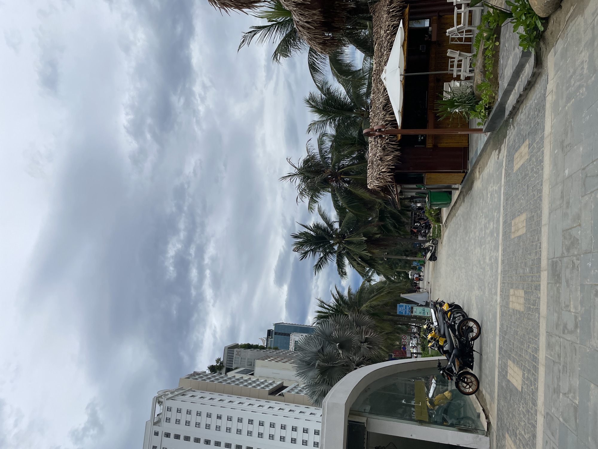 The image depicts a pathway lined with palm trees, leading towards a beach or coastal area in Da Nang, Vietnam. On the left side of the pathway, a modern, white multi-story building stands tall, with numerous windows visible. Adjacent to the building is a curved, glass-fronted structure. Several motorcycles are parked along the pathway in front of this structure. Further down the path, a series of small, thatched-roof huts are situated, one of which appears to be a bar or food stall with a white umbrella providing shade over outdoor seating. In the background, more palm trees and other buildings can be seen. The sky is overcast with heavy clouds. A few people are walking in the distance, and other motorcycles are parked further along the path. The pathway itself is paved with stone tiles.
