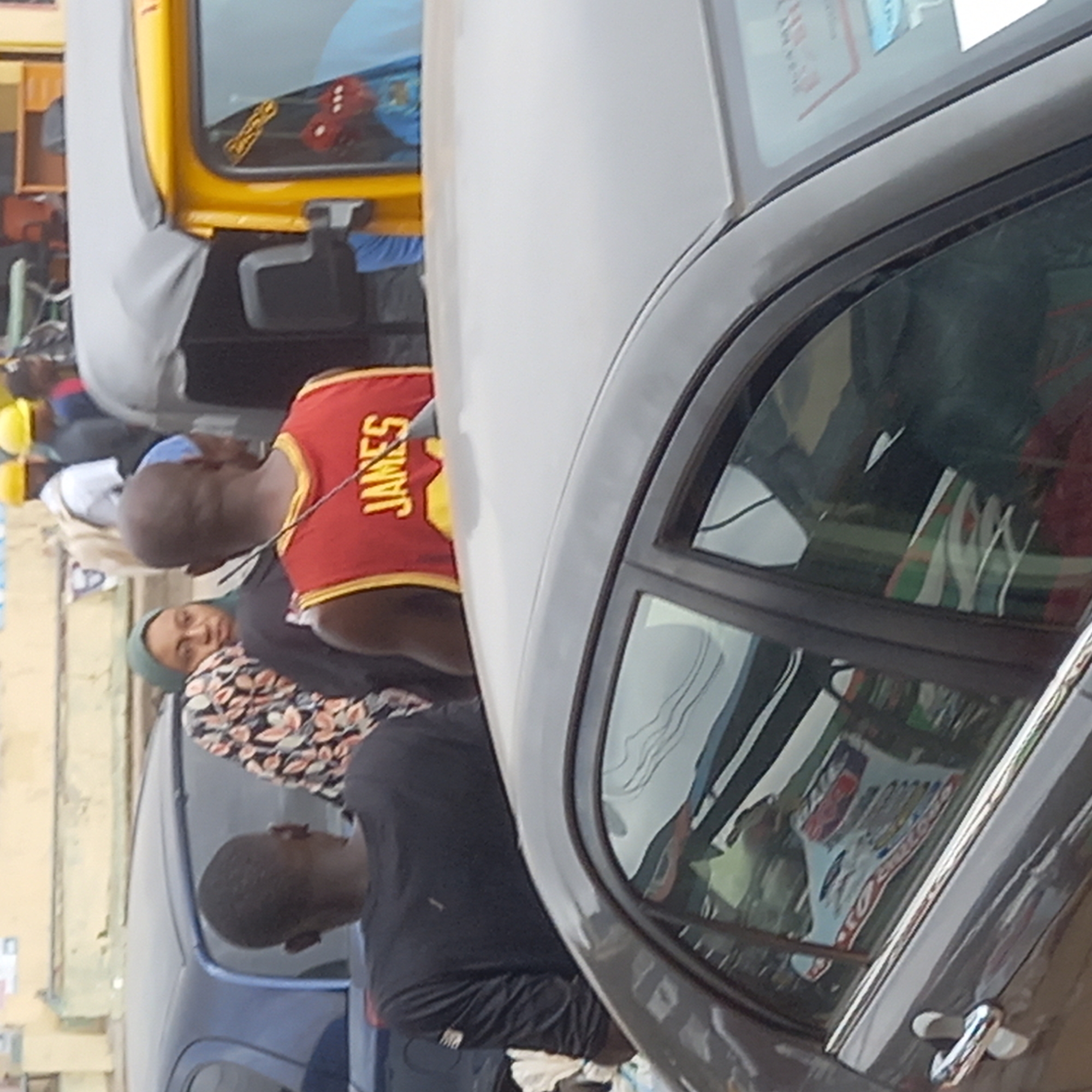 The image shows an outdoor scene in Dadi Riba, Nigeria. There are multiple individuals present. One man is wearing a red and gold "JAMES" basketball jersey. A woman in a headscarf and floral patterned shirt stands nearby. Another man is wearing a black shirt. The scene features a yellow three-wheeled vehicle (keke) and a dark grey car. The reflection in the car window shows a poster with red and white text. Other buildings are visible in the background.