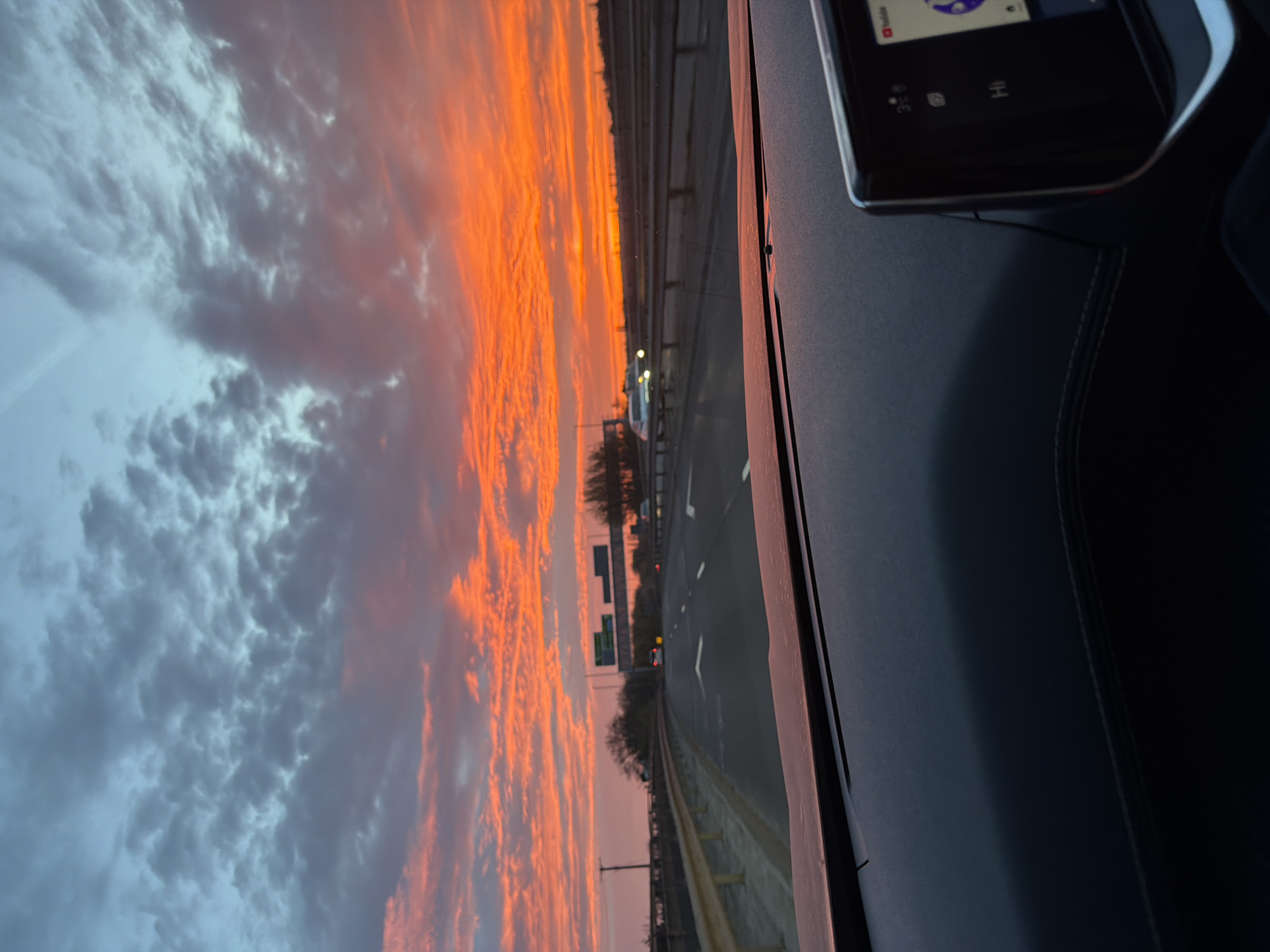 The image depicts a view from inside a car driving on a highway, likely in Garden City, United Kingdom, given the request. The highway is visible through the windshield with multiple lanes and guardrails. The sky is filled with a dramatic sunset, with streaks of orange and grey clouds. Part of the car's dashboard is visible, including a screen displaying a user interface and a portion of the seating area. There are no people visible. The action is the movement of the car on the highway. The interaction is the driver observing the sunset while driving. The environment is a combination of urban highway infrastructure and a dramatic sky.