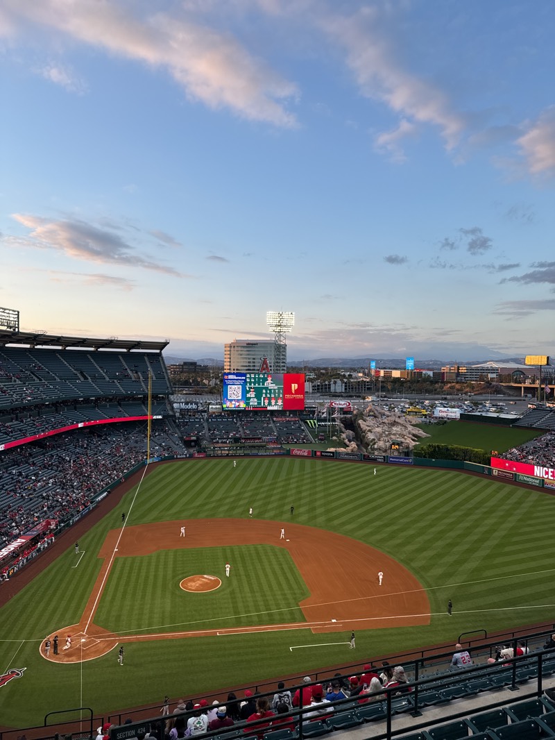 The image captures a high-angle view of a baseball game in progress at Angel Stadium in Anaheim, California, United States. The field is visible, with players in uniform positioned on the diamond and in the outfield. Spectators fill a significant portion of the stadium seating. A scoreboard displaying information is visible in the background, along with advertisements. Outside the stadium, buildings and parking lots are present, with mountains visible in the distance. The sky is partly cloudy.