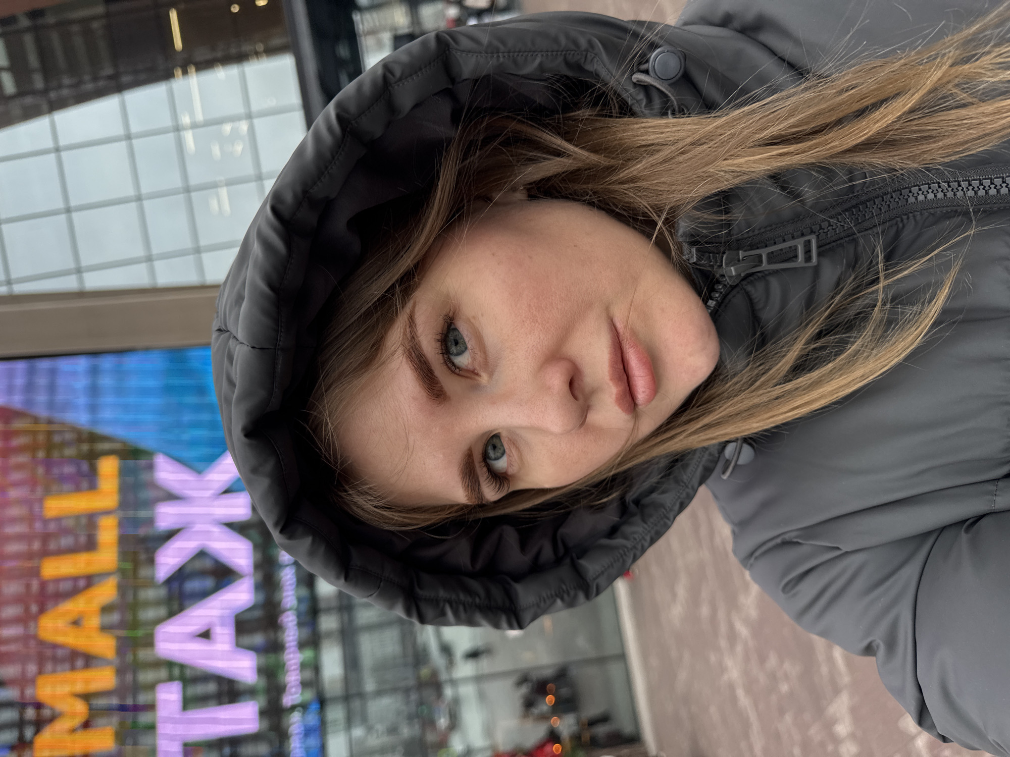 A young woman with long blonde hair and blue eyes is wearing a gray hooded puffer jacket. She is looking at the camera with a slight smirk. The backdrop includes a building with a sign in Russian that reads "MALL TAK," which is likely referring to the city of Perm, Russia. Reflections in the building's glass façade suggest it is an outdoor setting.