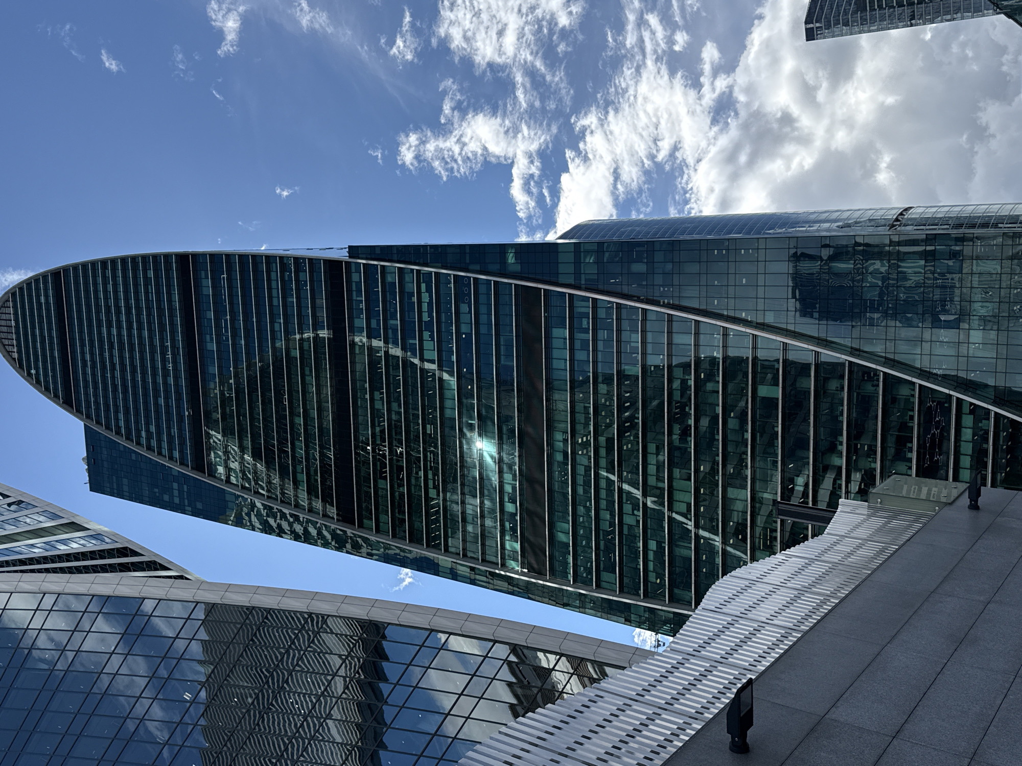 The image is a low-angle shot of modern skyscrapers in Moscow, Russia. The dominant structure is a uniquely curved, glass-clad building that tapers towards its apex, reflecting the bright blue sky and scattered white clouds. To its left, another glass skyscraper stands, its facade also mirroring the sky and clouds, with visible horizontal banding. In the foreground, a dark gray stone wall runs horizontally across the lower part of the frame, topped by a white, perforated metal fence with a wavy pattern. Two black light fixtures are attached to the stone wall. The overall scene is one of architectural grandeur and urban development, bathed in natural daylight.