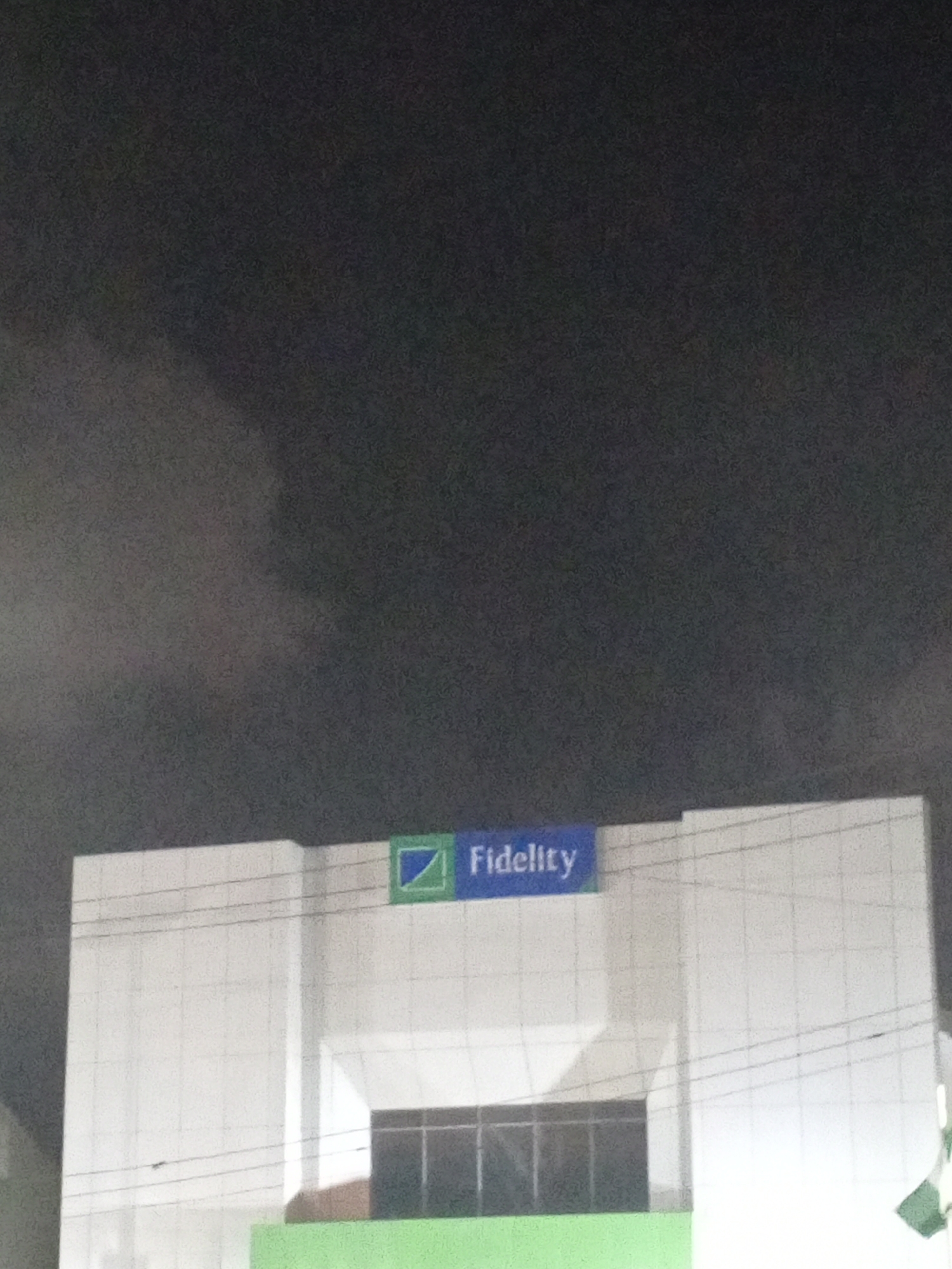 The image shows the Fidelity Bank building at night in Lagos, Nigeria. The building is white with a green base and a large window. The "Fidelity" logo is displayed in blue and green on top of the building. The sky is dark and cloudy. There are no people visible in the image.