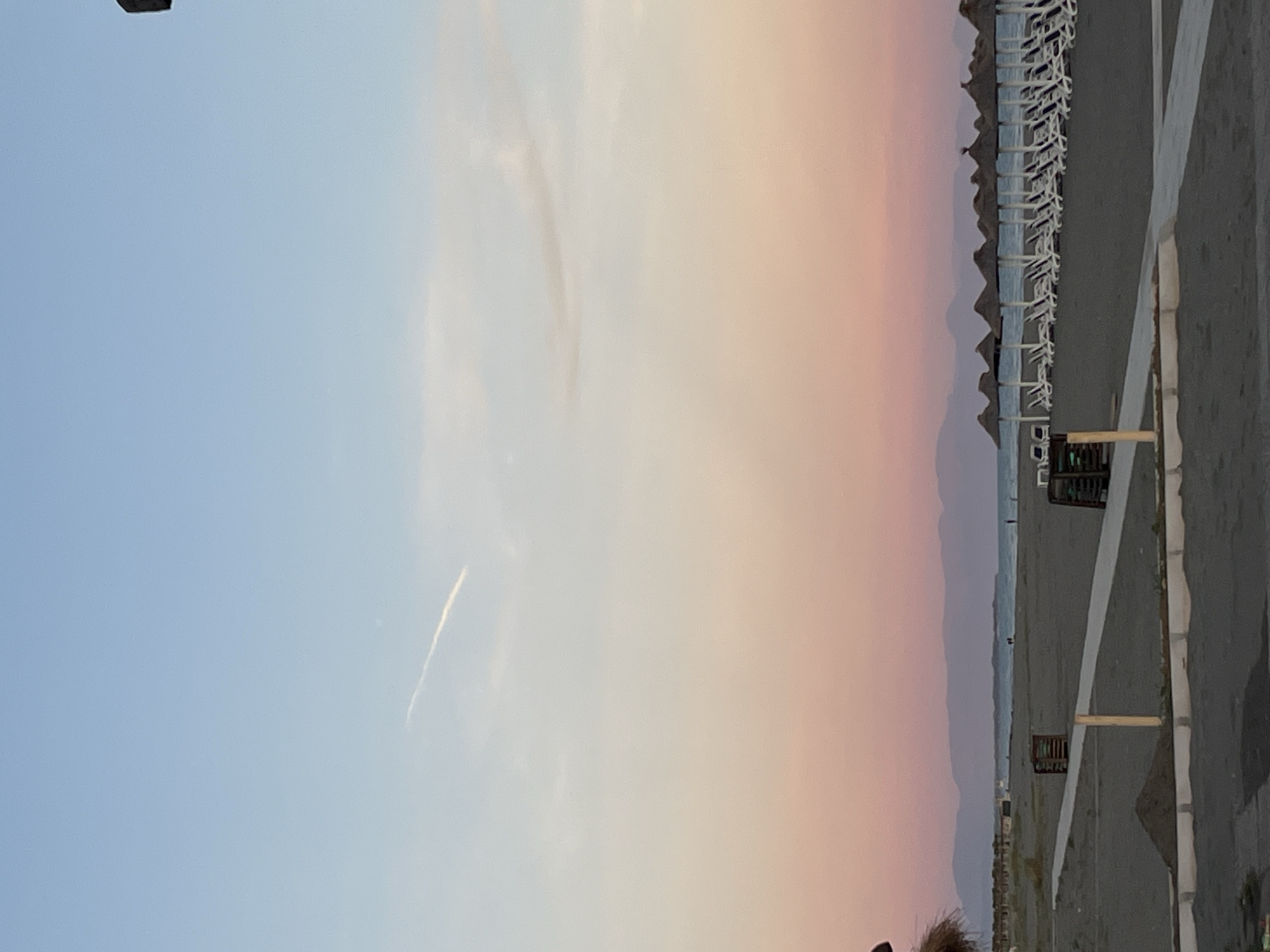The image depicts a serene beach scene at sunset in Pistula, Montenegro. The sky displays a gradient of colors, transitioning from a soft blue at the top to warm pink and orange hues near the horizon. A faint airplane contrail streaks across the upper portion of the sky. In the foreground, dark sand covers the beach, with a pathway made of concrete or paving stones. Along the pathway, there are two wooden structures, possibly serving as planters or bins. A small pile of sand is visible near the pathway. Further back, a row of thatched umbrellas stands over empty lounge chairs arranged on the sand, facing the calm sea. In the distance, a line of mountains creates a silhouette against the colorful sky, and a few indistinct figures can be seen walking near the water's edge. The overall impression is one of tranquility and the quiet beauty of a coastal evening.