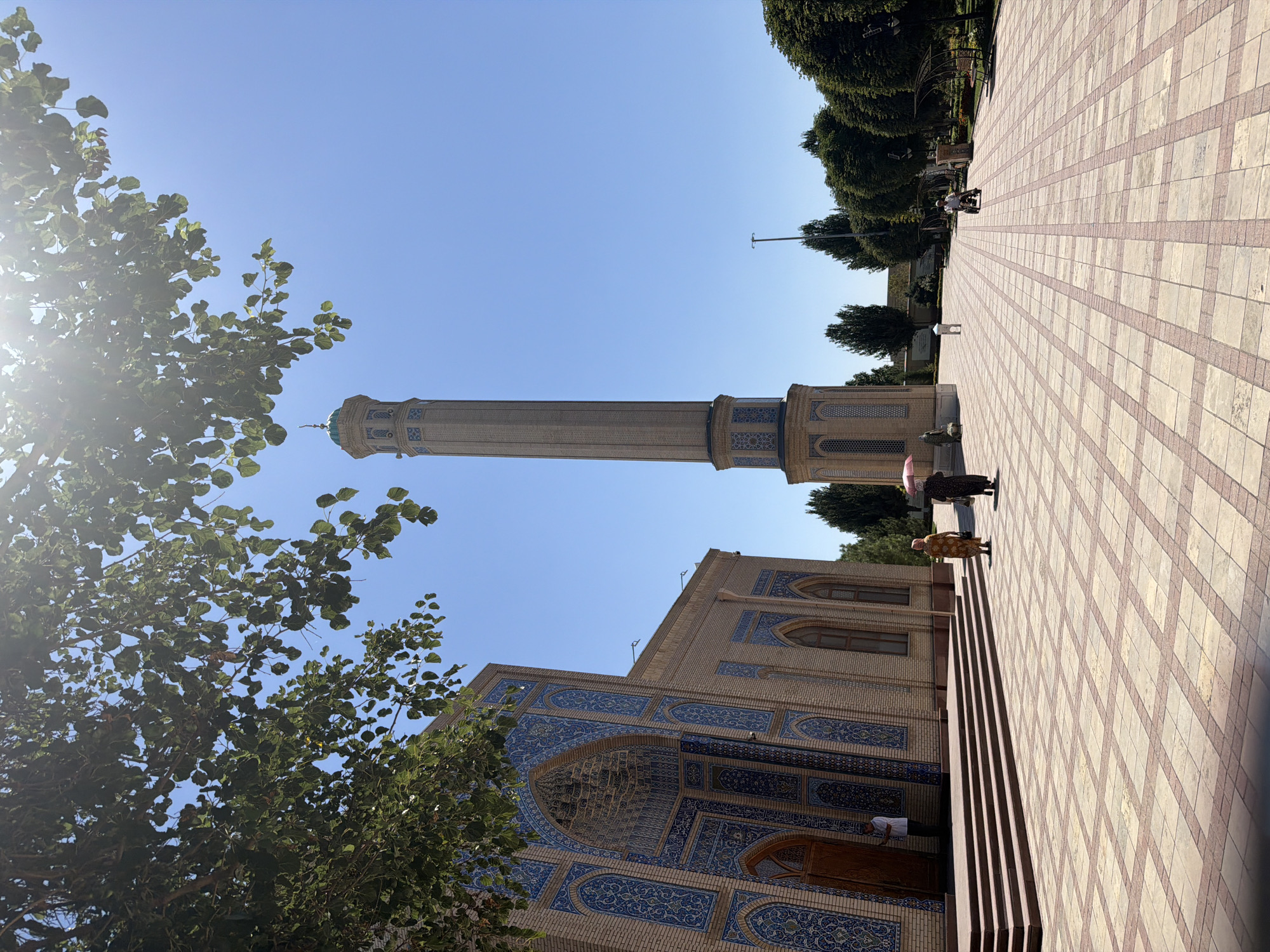 This is an outdoor, eye-level, full shot captured on a sunny day in the city of Zangiata, Uzbekistan. The image features a mosque with a tall minaret to the right. The mosque's facade is adorned with intricate blue and beige tilework, and a wooden door is visible. In front of the mosque, a tiled courtyard stretches out, with a series of steps leading up to the entrance. Two women, one in a yellow dress and the other in a dark dress, walk across the courtyard, with the woman in the dark dress holding a pink umbrella. The minaret is slender and cylindrical, decorated with blue tile patterns. Further into the courtyard, trees are planted in neat rows, and benches are visible. A person in a white shirt is operating a small motorized scooter in the middle ground. The sky is clear and blue, with sunlight filtering through the leaves of a tree in the upper left corner of the frame.
