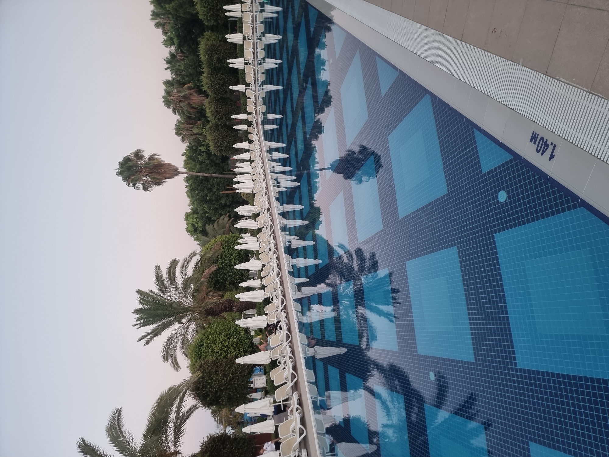 The image shows a swimming pool area at a resort in Manavgat, Turkey, possibly during twilight. The pool itself has a checkered bottom with blue and darker blue squares. The water reflects the surrounding palm trees, white lounge chairs, and closed white umbrellas, creating a symmetrical pattern. A depth marker indicating "1.40 M" is visible along the edge of the pool. In the background, a line of lounge chairs and umbrellas are neatly arranged. Lush green trees and palm trees dominate the landscape behind the pool area. The sky is a pale, clear gradient, suggesting either early morning or late evening. There are no people clearly visible or engaged in any activity within the frame, though a figure is partially obscured near a chair in the background.